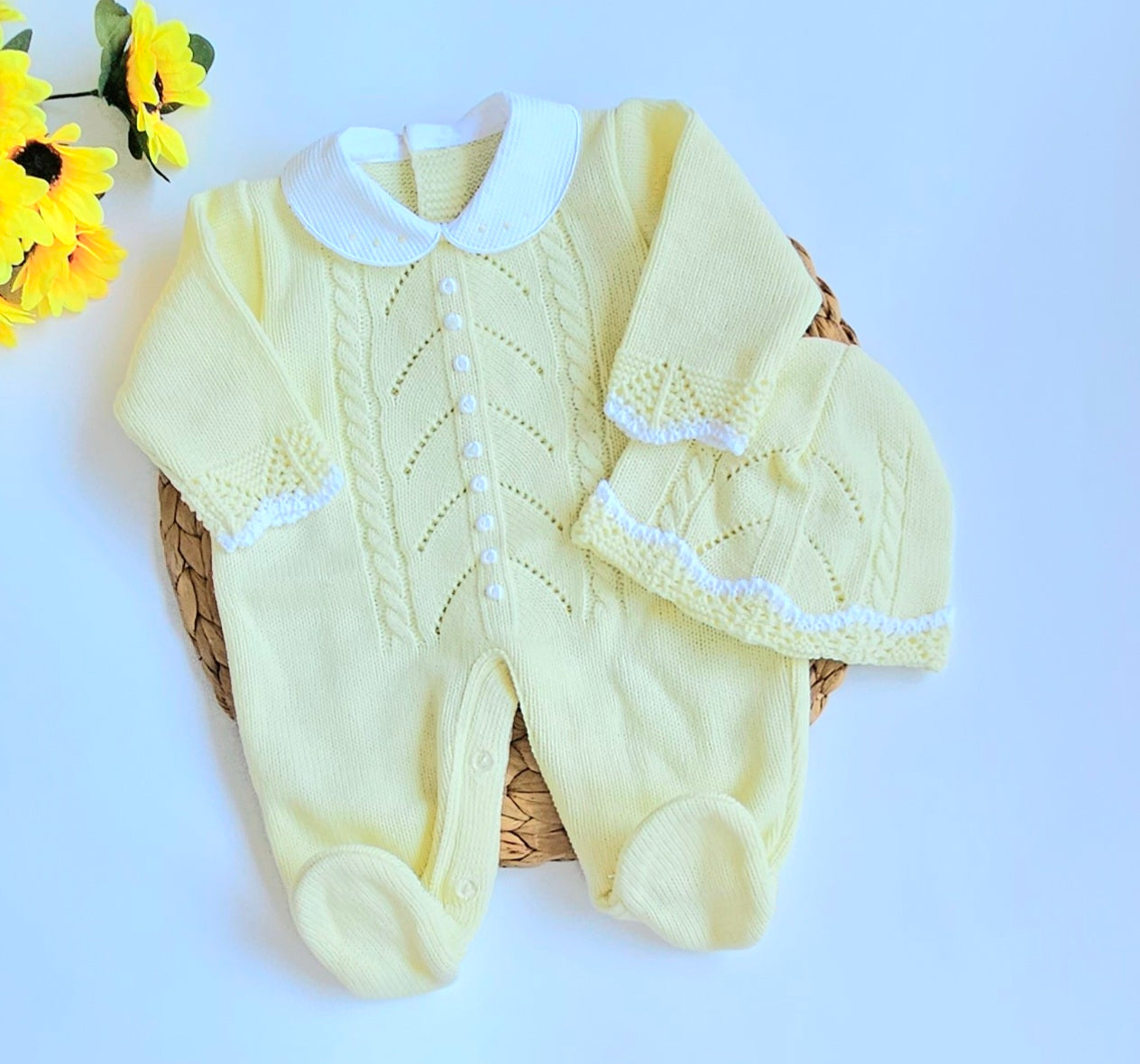 A yellow knitted baby romper with handmade embroidery, accompanied by a matching hat, displayed on a basket.