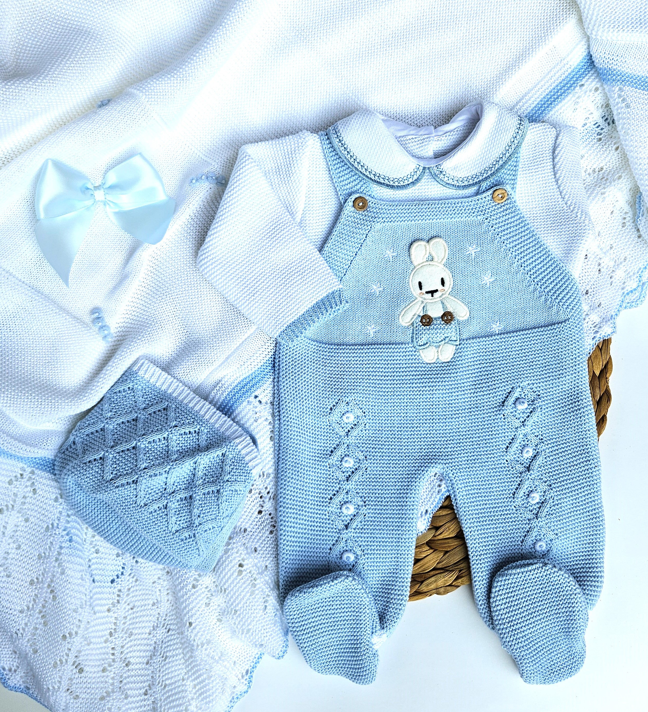 A set of light blue knitted baby clothes with a cute bunny face on the front, accompanied by a white blanket with blue details, presented together.