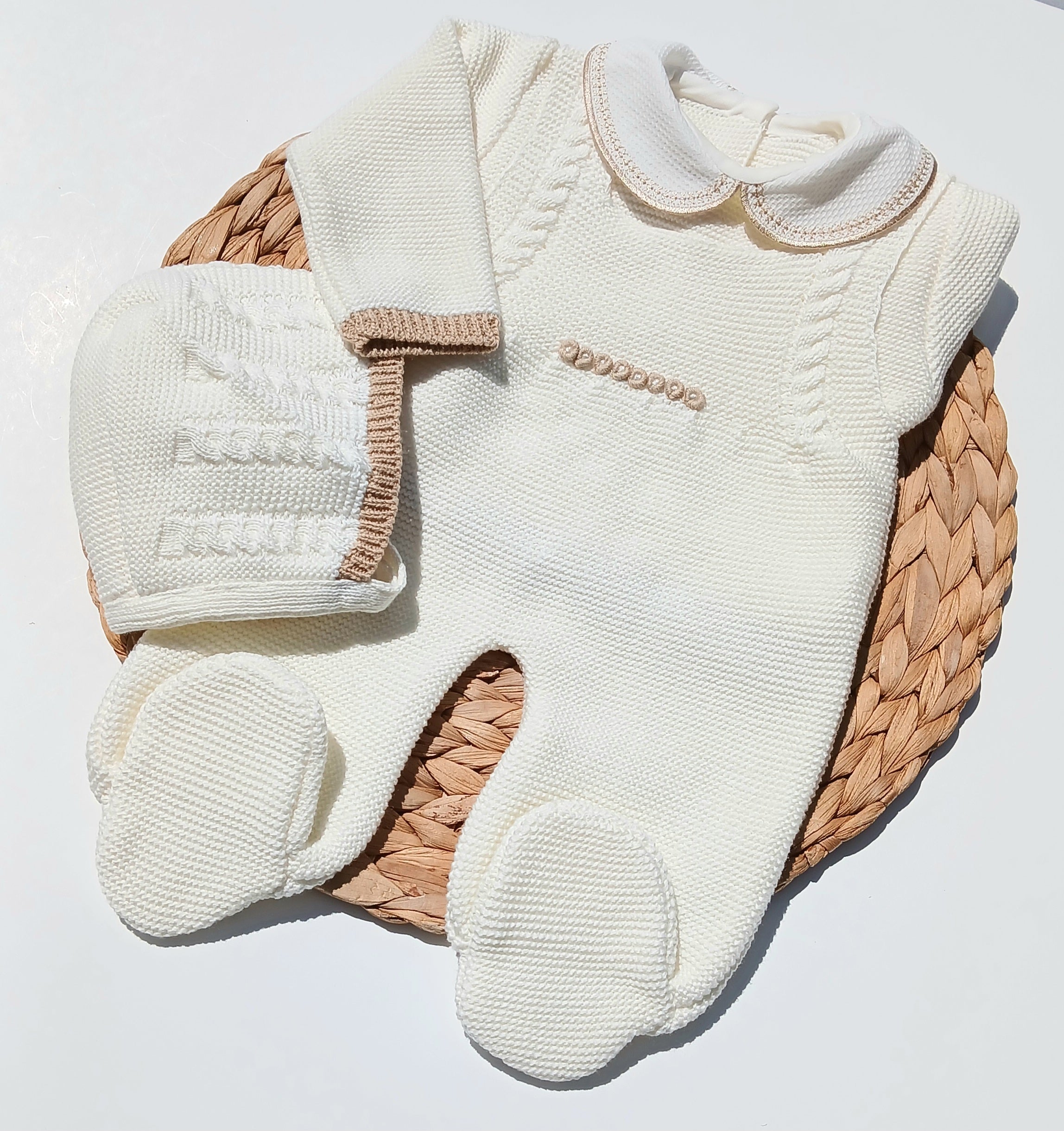 Baby outfit with white knitted romper, hat, and booties on a woven basket. newborn neutral outfit Cream-colored baby outfit with matching hat and booties on a woven basket against a light background