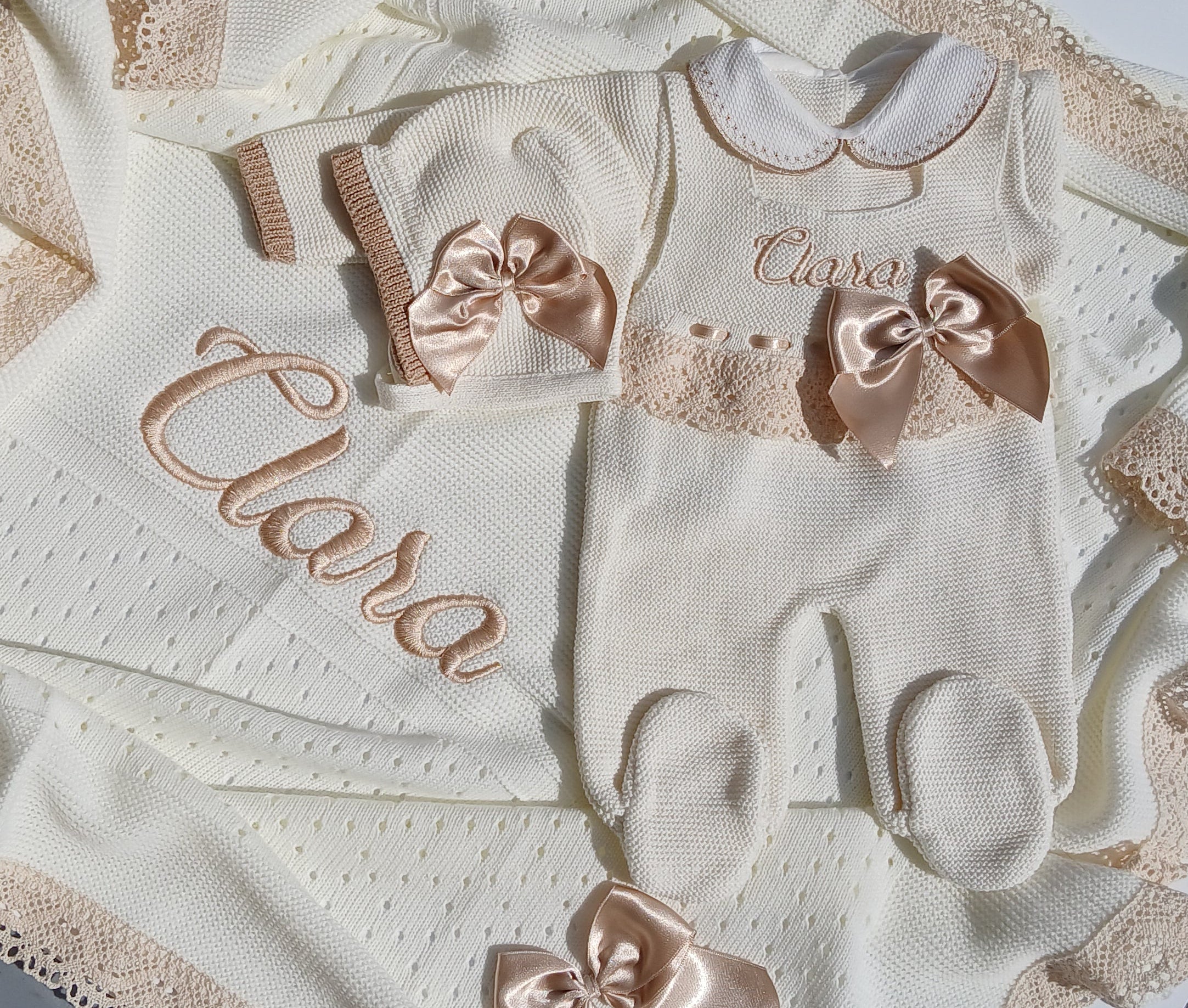 A baby clothing set laid flat on a white surface, including a cream bodysuit with lace trim, matching booties, a small bonnet, and a blanket with embroidered script and lace edges. A person's hand is visible in the top right corner.