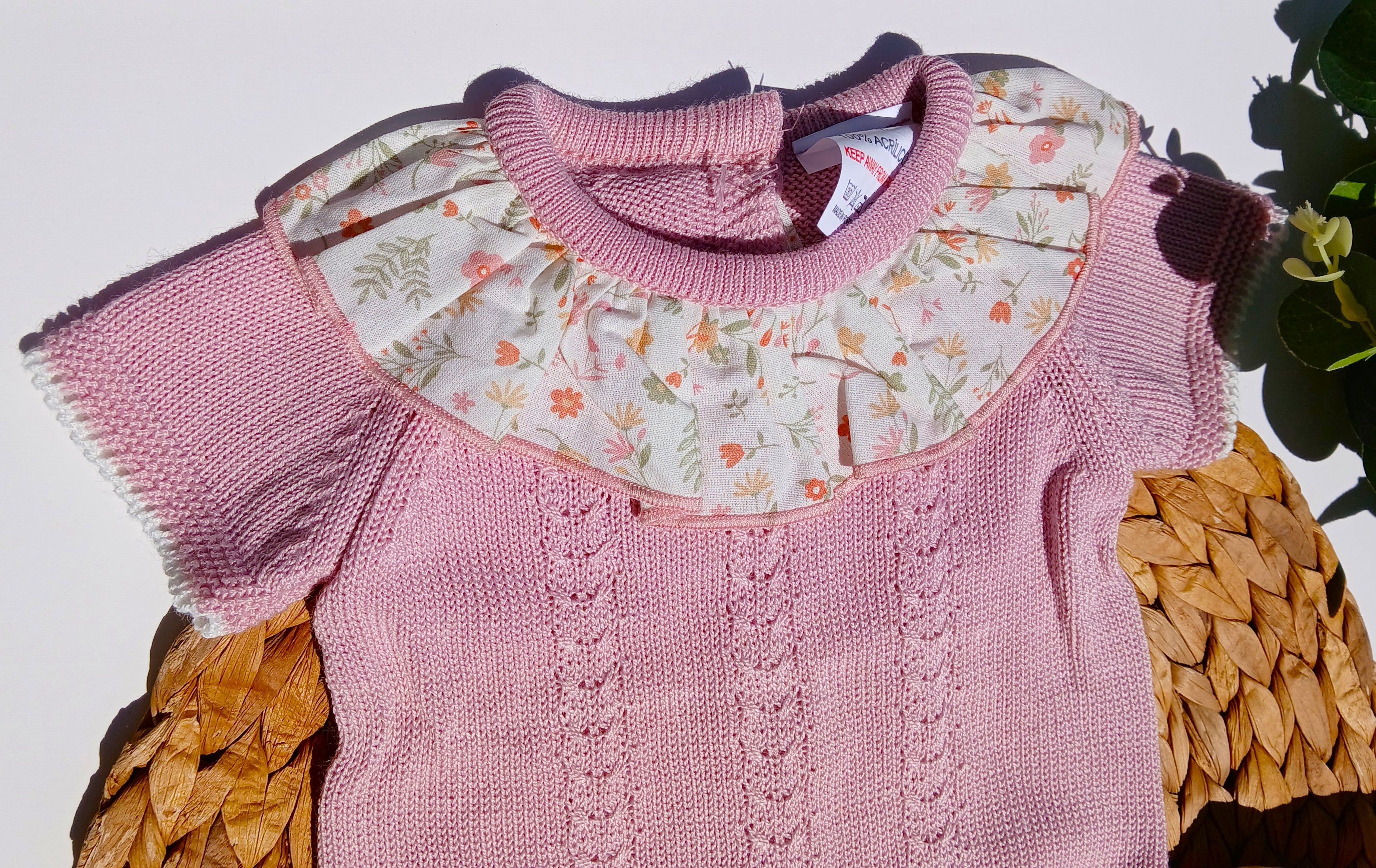 Pink knitted baby outfit with floral collar on a textured surface