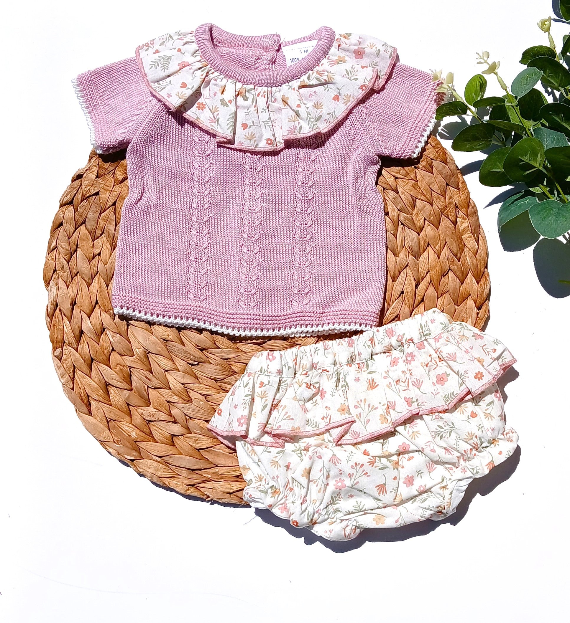 Pink baby sweater and floral bloomers on a woven mat with a plant in the background