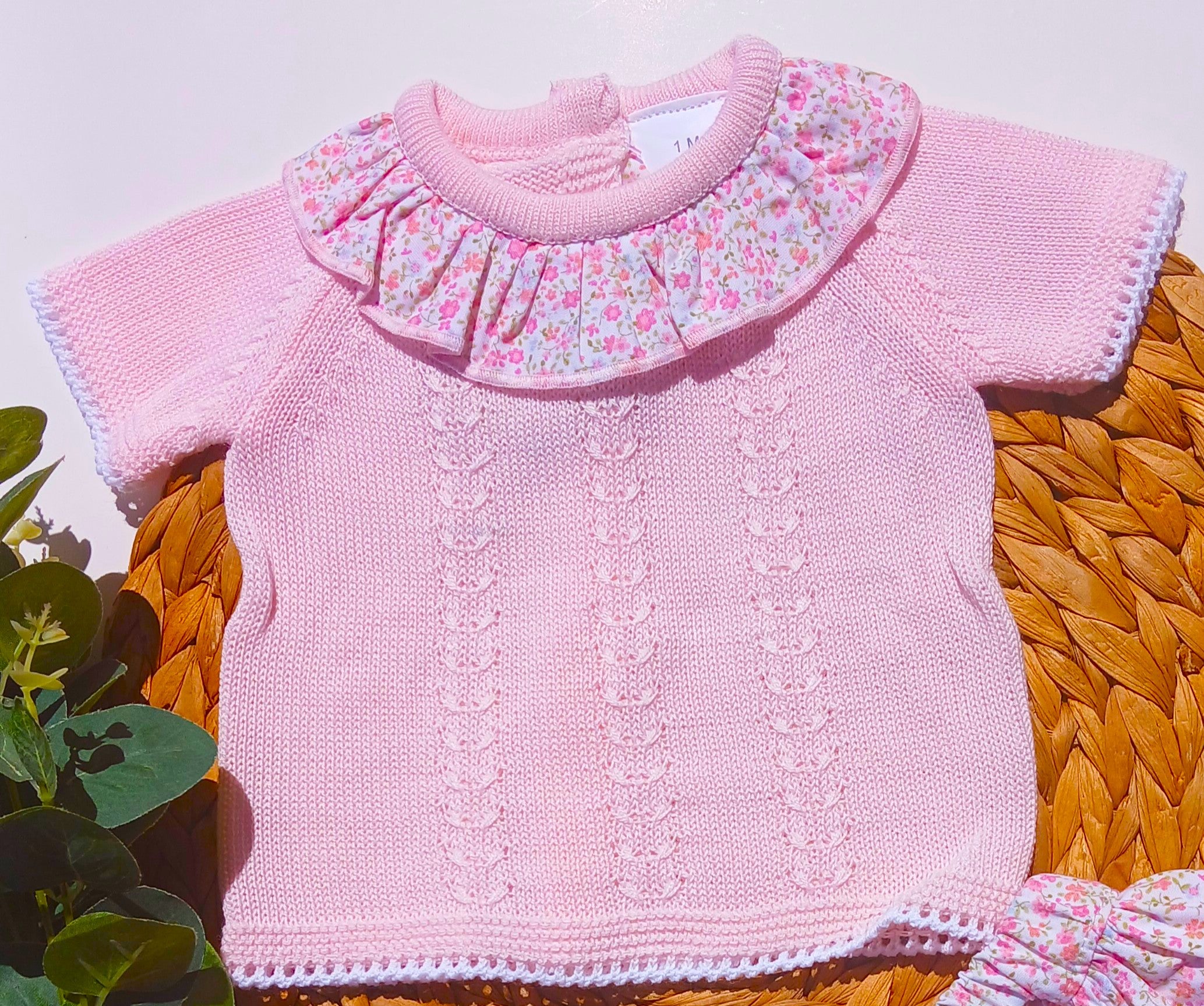 Pink knitted baby sweater with floral collar on a woven surface