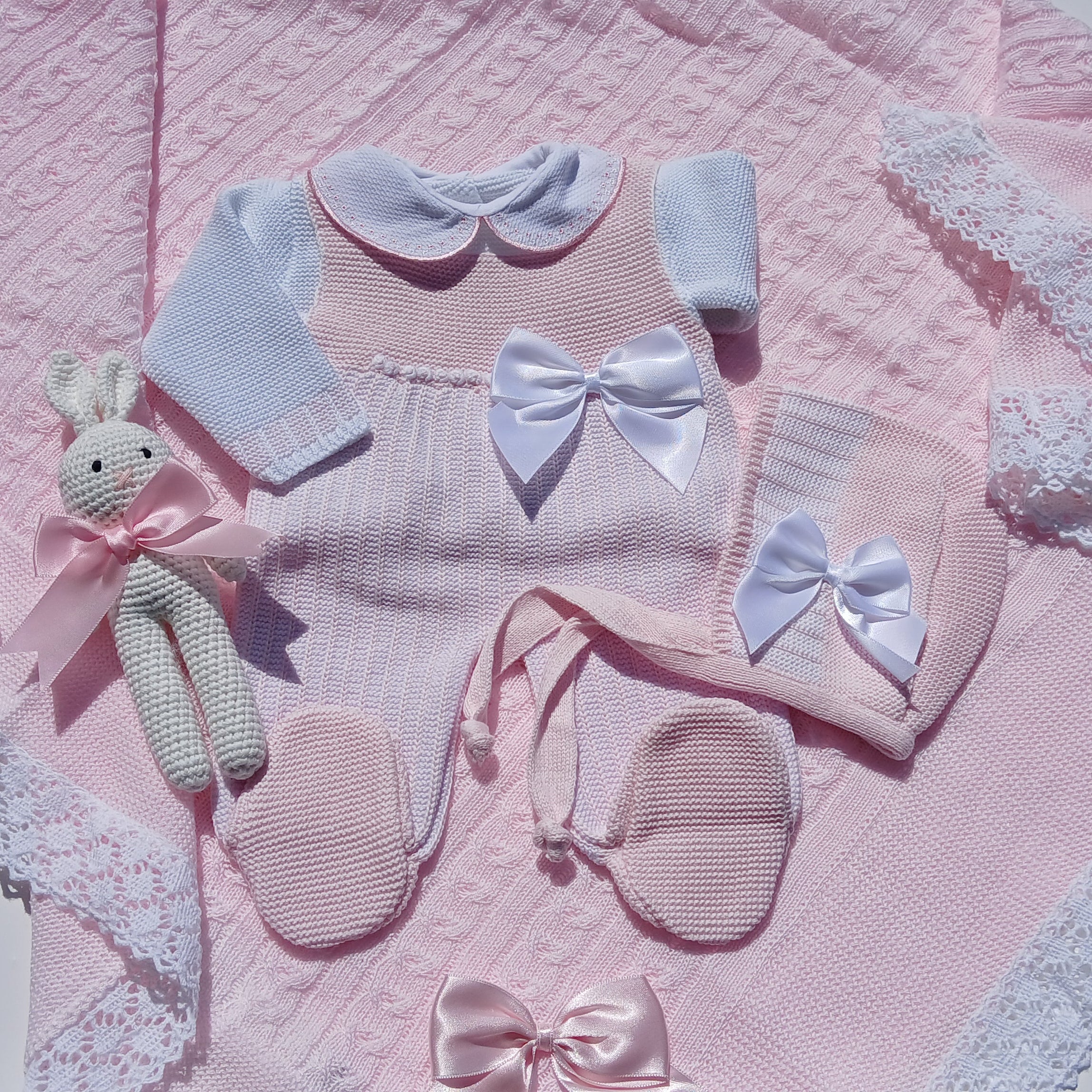Collection of baby clothes and accessories on a pink blanket with white lace trim.