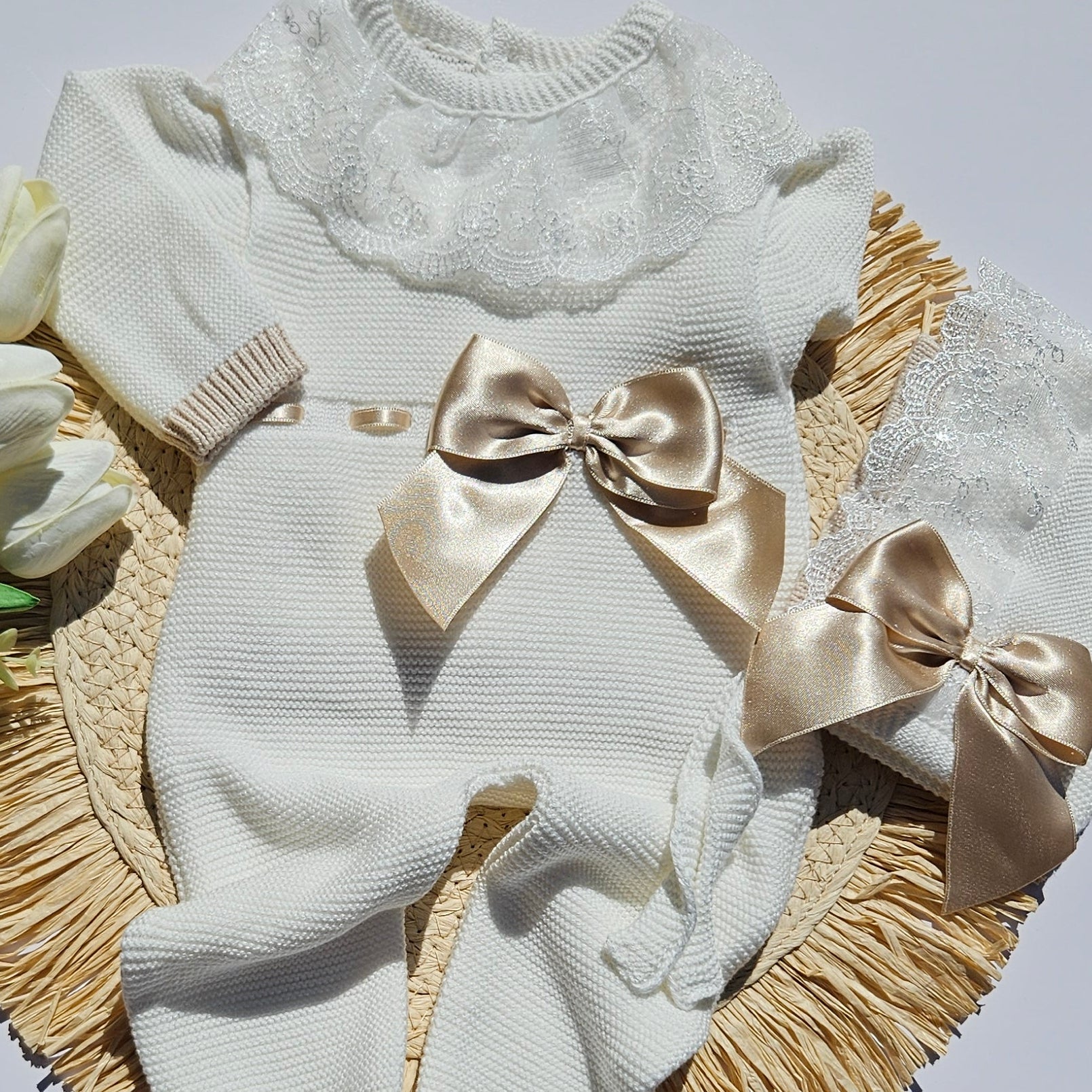 A baby outfit set including a cream knit romper, matching hat, and white lace bib, styled on a straw hat with beige ribbons and white tulips, displayed on a white board.
