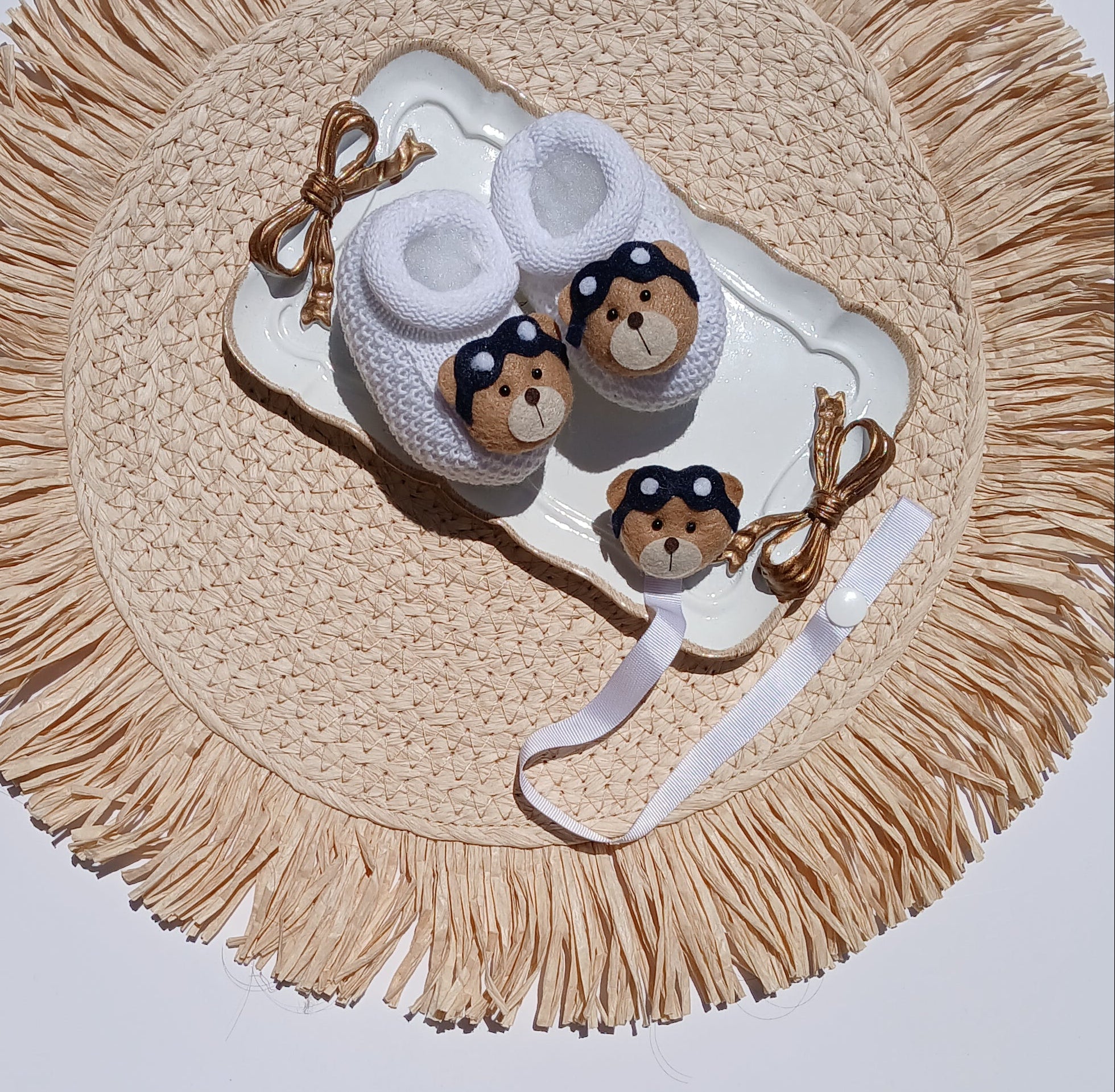 A pair of white knit baby booties with brown and black patterned socks and a matching white headband, arranged on a round woven straw placemat.