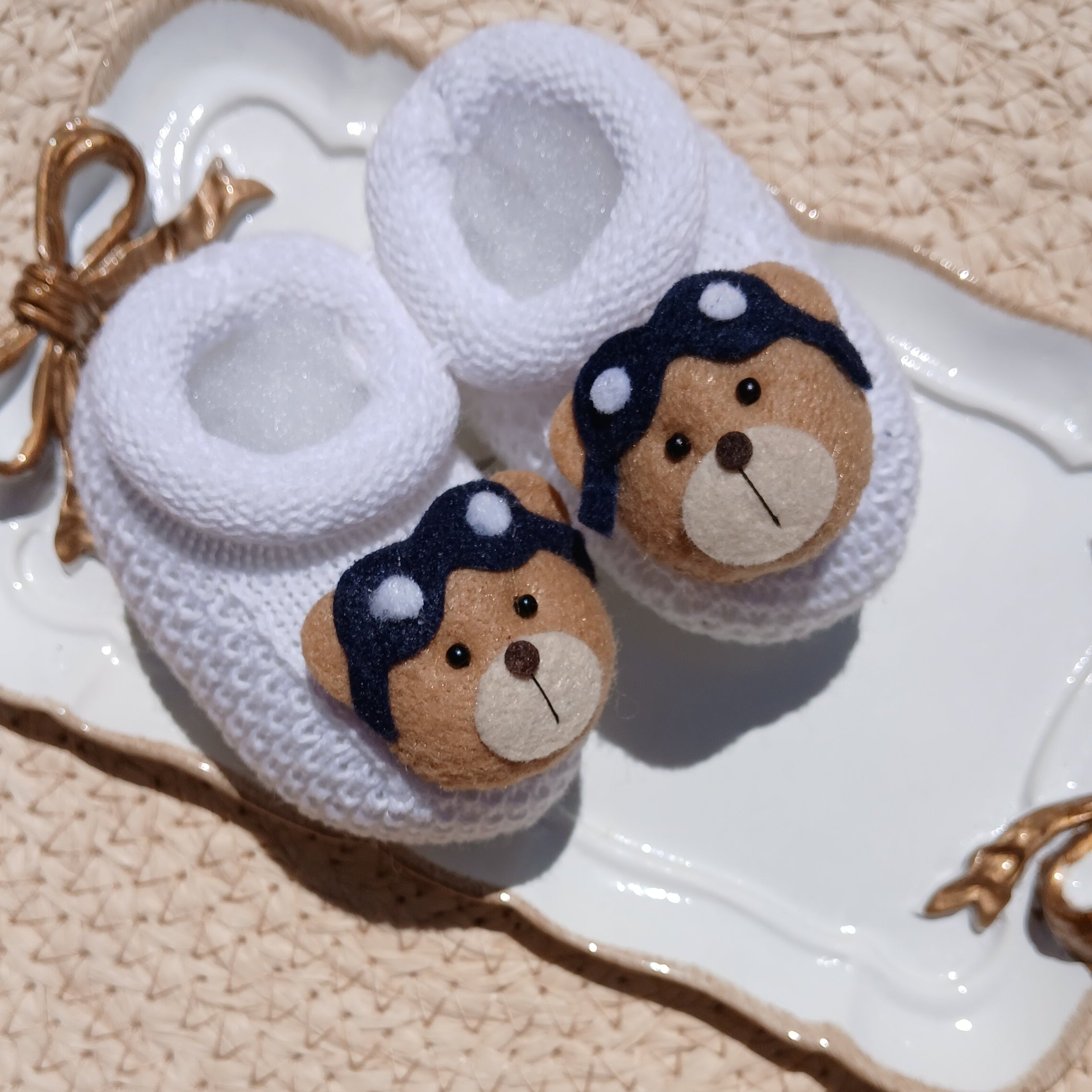 A pair of white knit baby aviator booties with embroidered teddy bear faces and navy bows, displayed on a white ceramic dish over a beige woven mat.