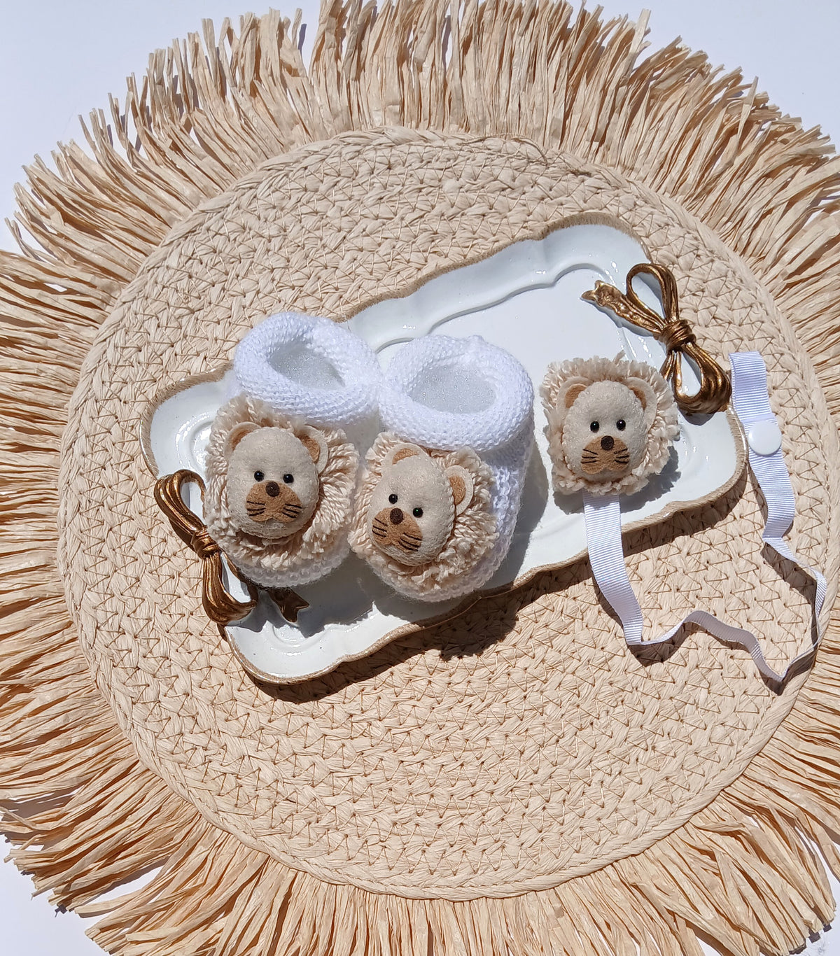 A set of baby accessories including a pair of white knit booties with lion face details and a matching white bonnet, displayed on a white tray over a woven straw placemat.