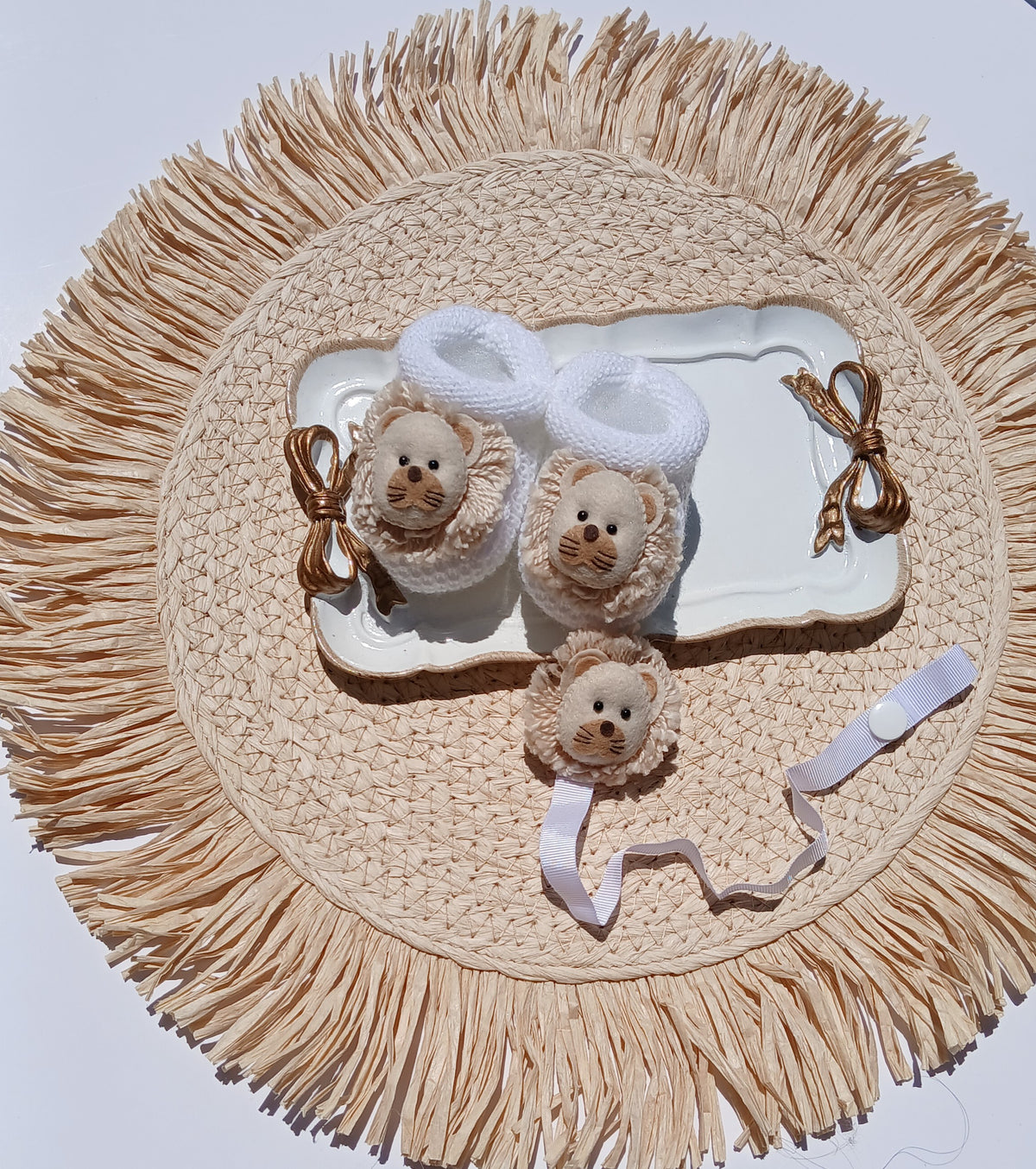 A baby accessory set featuring a pair of white knit booties with lion faces and brown ribbon bows, a matching lion head band, and a small cream-colored plush or fabric lion toy, all arranged on a white tray over a round woven straw placemat. Lion Pacifier chain
