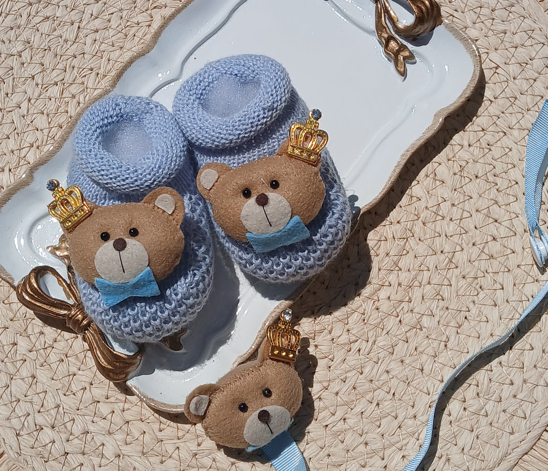 A pair of light blue knit baby booties with plush teddy bear faces and gold crown details, displayed on a white ceramic dish over a woven straw hat.