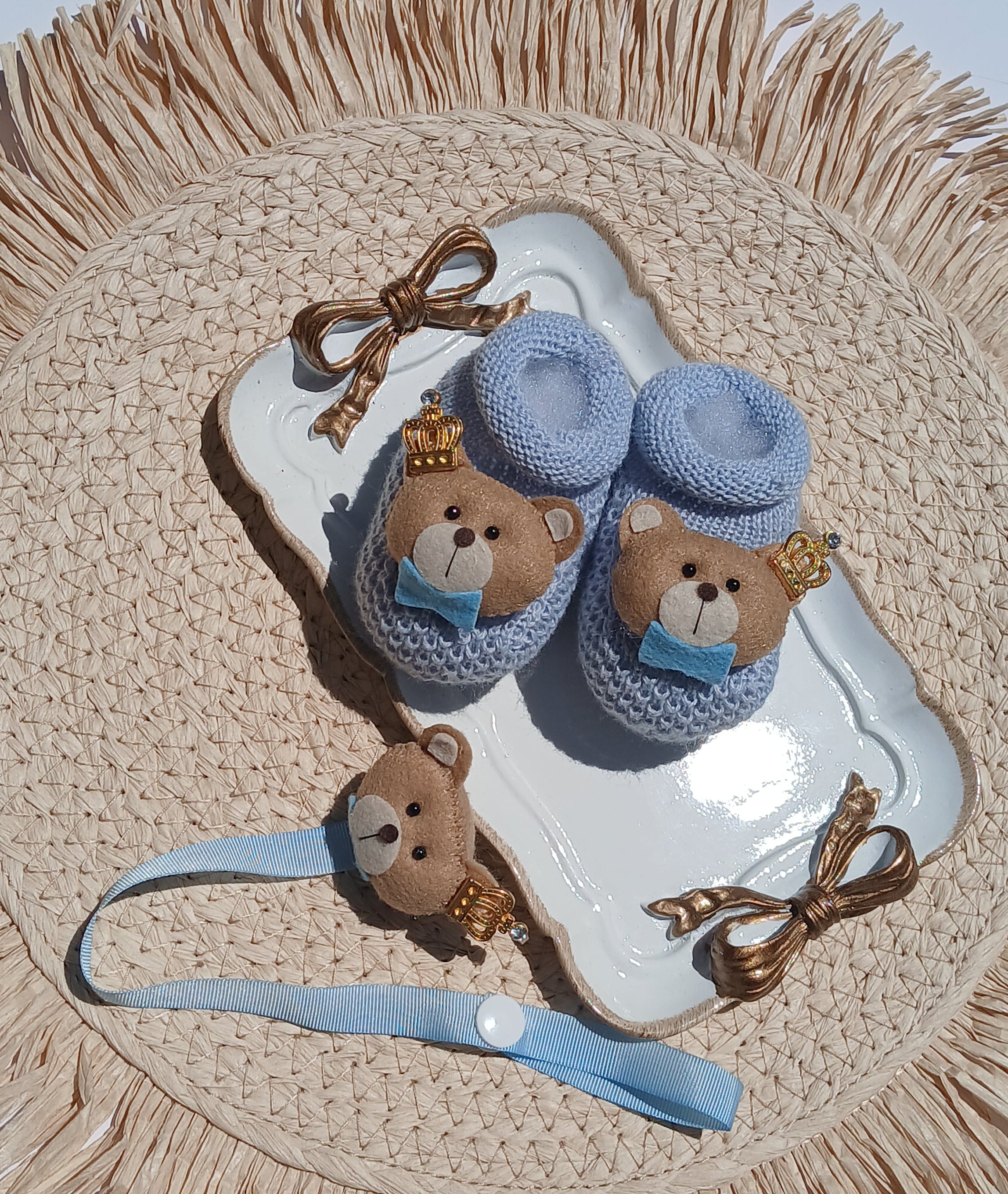 A baby accessory set featuring a pair of light blue knit booties with teddy bear faces, a matching blue and brown headband, and a small brown teddy bear hair clip, all arranged on a white tray over a woven straw placemat.