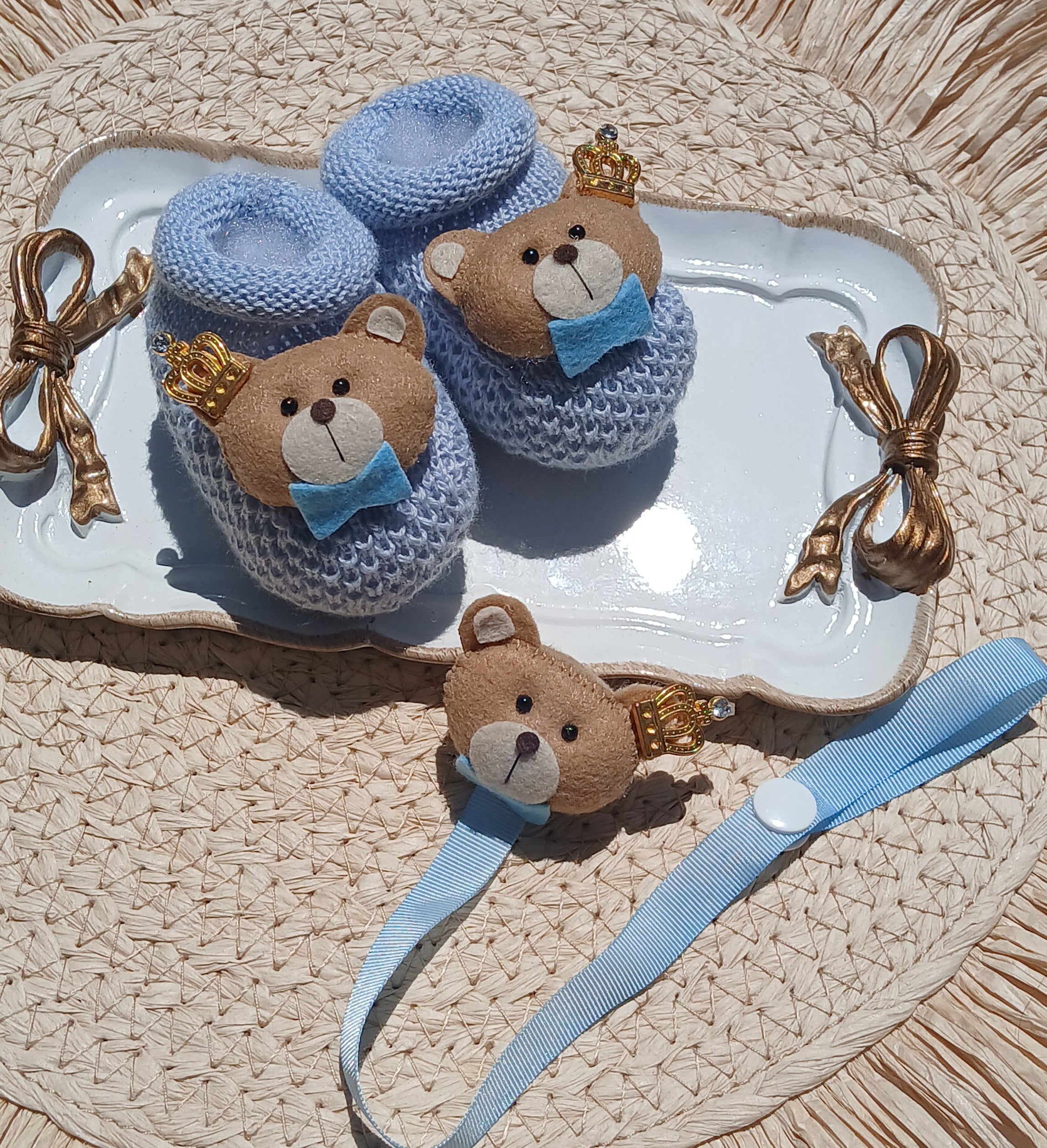 A styled flat-lay of a baby keepsake set featuring a light blue knitted baby blanket with a teddy bear face, a matching blue ribbon, and a small brown teddy bear charm, arranged on a white ceramic dish over a woven straw mat.