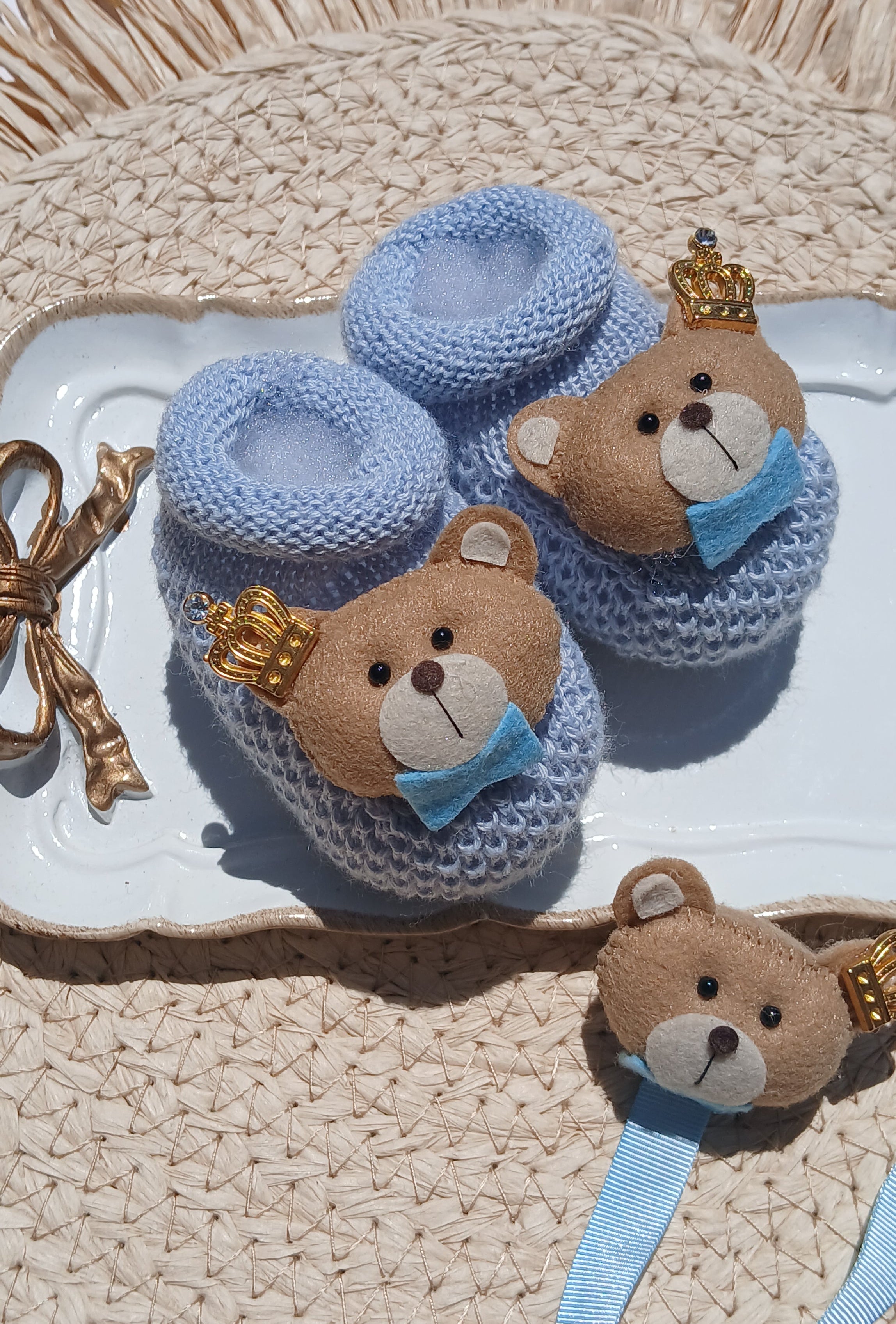 A pair of light blue knitted baby booties with plush teddy bear faces and gold bows, displayed on a white tray next to a woven straw hat.