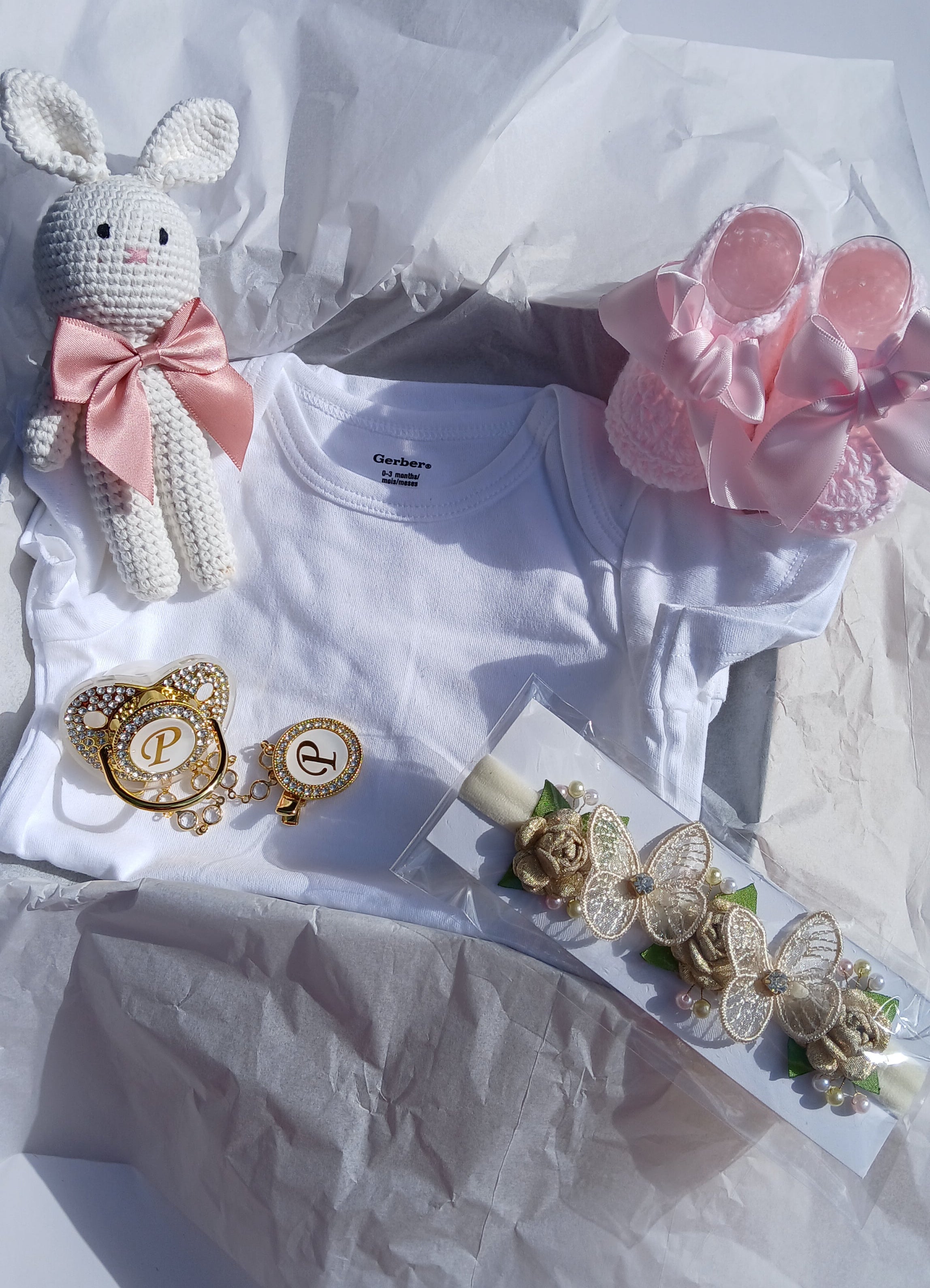 A baby gift set arranged on white tissue paper, featuring a white short-sleeve baby shirt, a white crocheted bunny with a pink bow, a pair of pink booties, gold-tone jewelry including a heart-shaped brooch and a floral headband, and a small white card.
