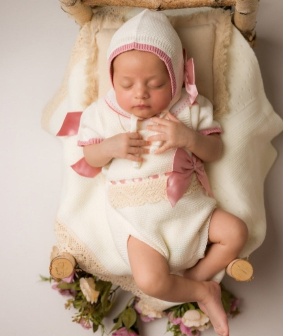 Newborn baby in a white outfit with pink accents lying in a wooden crib. Newborn baby in a knit outfit and bonnet lying on a wooden crib with flowers around.