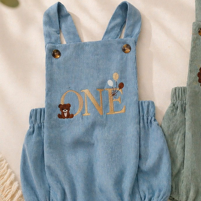 Two baby rompers with 'ONE' embroidery on a soft surface.