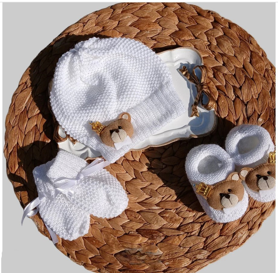 White knit baby hat and mittens with small teddy bear face details, displayed on a woven brown basket with matching booties.