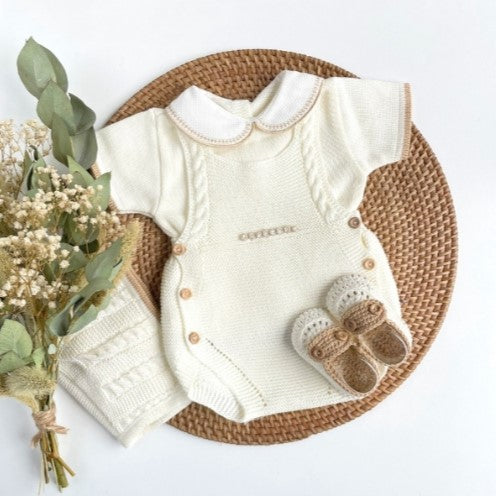 Baby outfit with pearl Ivory knitted cardigan, pants, and shoes on a woven mat with flowers. newborn summer outfit