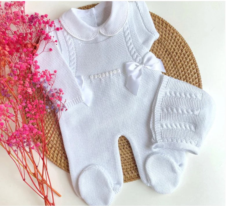 White knitted baby romper with a bow on a woven mat with pink flowers.
