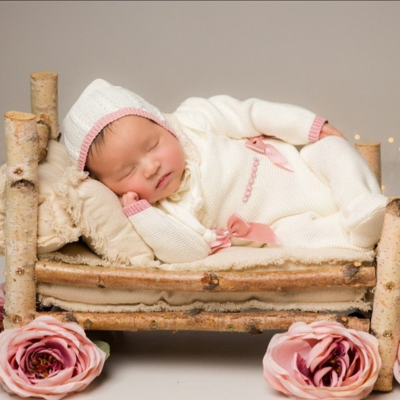 Newborn baby in a white outfit with pink accents lying on a small wooden bed surrounded by pink flowers. , newborn knit going home outfit, newborn baby girl coming home knit outfit