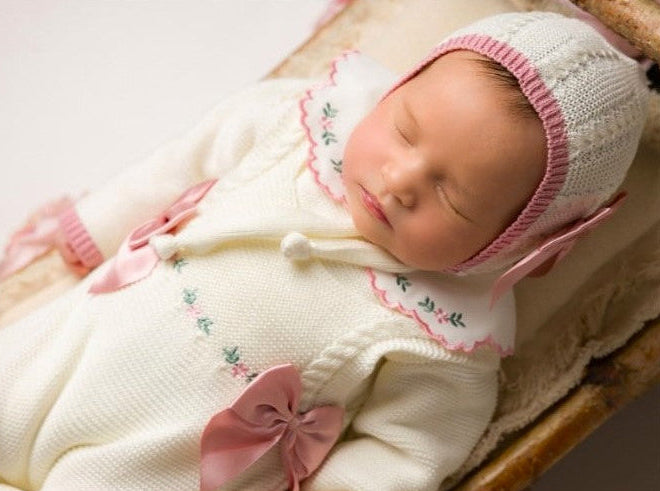 newborn girl knit going home outfit with flowers embroidered details
Ivory color with Old rose / mauve bow cetin. Baby girl knit coming home outfit in Ivory and Old rose details, collar with handmade flowers Newborn baby in a white outfit with pink accents, including a hat and bow, sleeping peacefully.