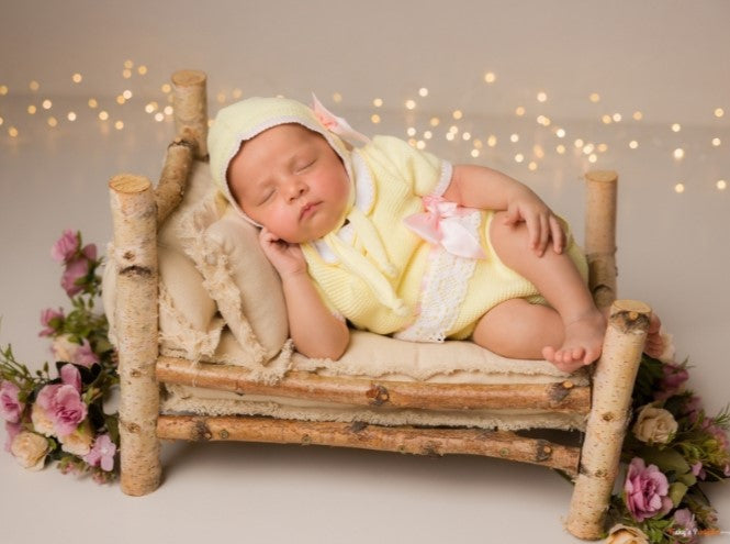 Newborn baby in a yellow outfit and bonnet lying on a small wooden bed with flowers and lights in the background.