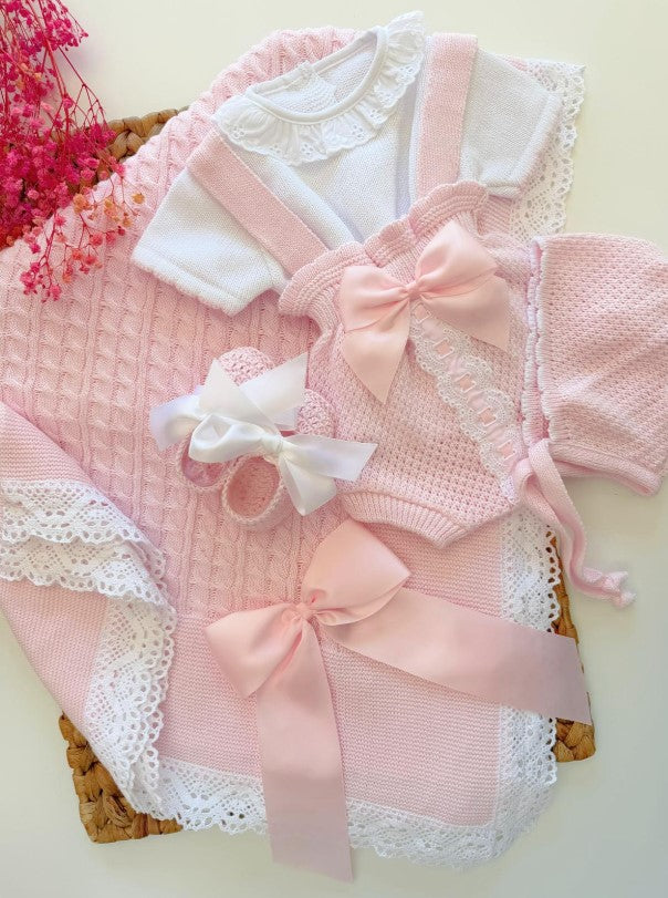 A pink and white baby girl's clothing set laid flat on a light background, featuring a white short-sleeve top with lace trim, a pink knit romper with a large pink bow, a matching pink knit cardigan with white lace trim, a small white bow hair clip, and a pink knitted hat.