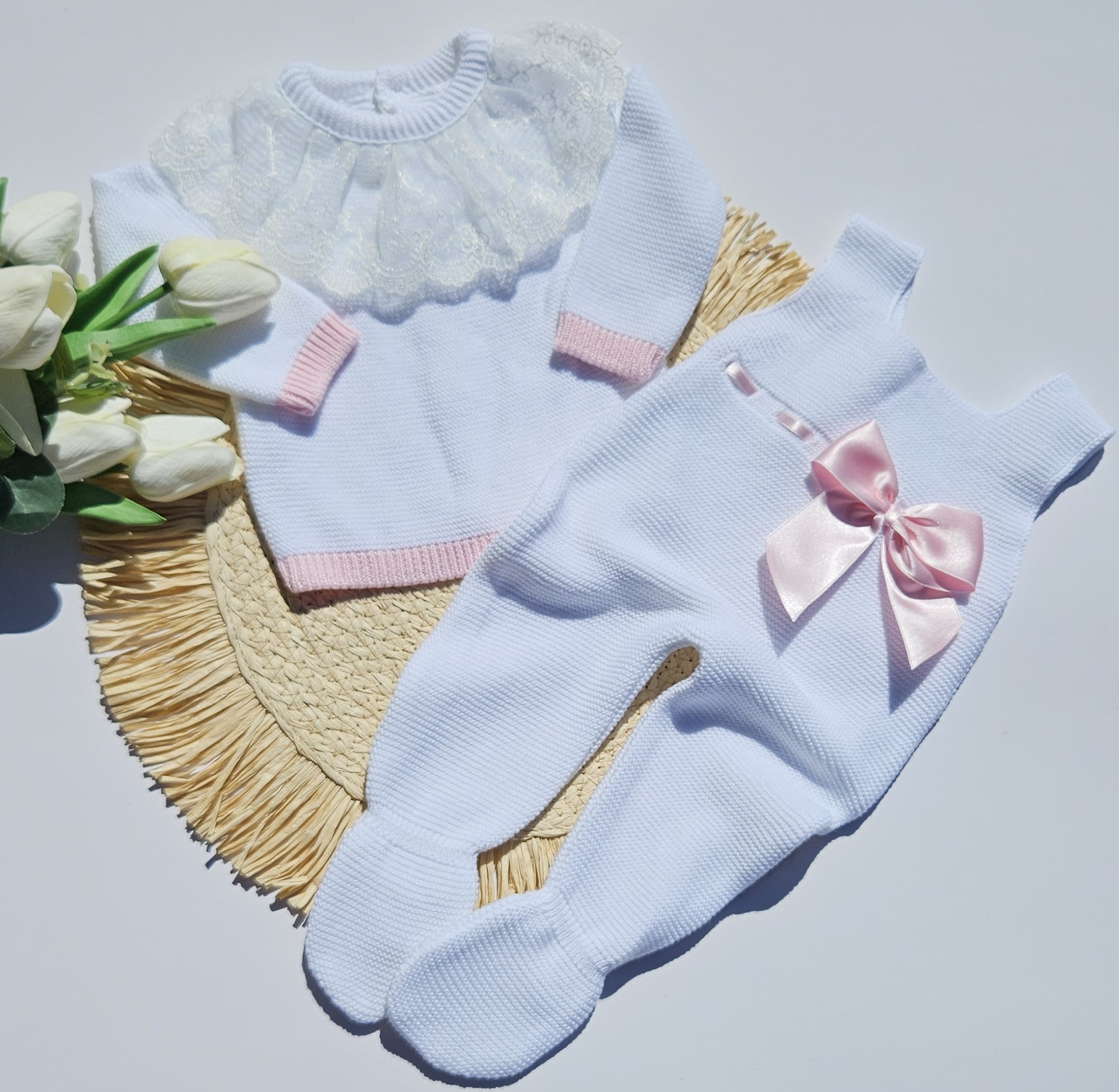 A baby footed romper in pale blue knit with a pink ribbon bow, laid flat on a woven mat with a matching pale blue top and white lace collar, next to white tulips.