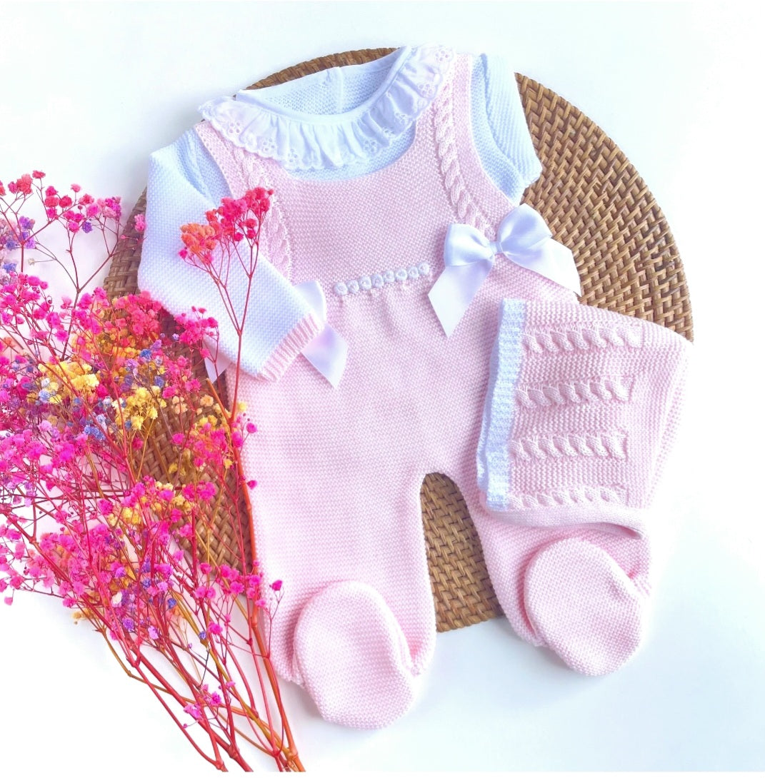 A newborn baby outfit set in pink and white, including a long-sleeve onesie, footies, and a matching bonnet, presented on a wicker surface with pink and red dried flowers to the side.