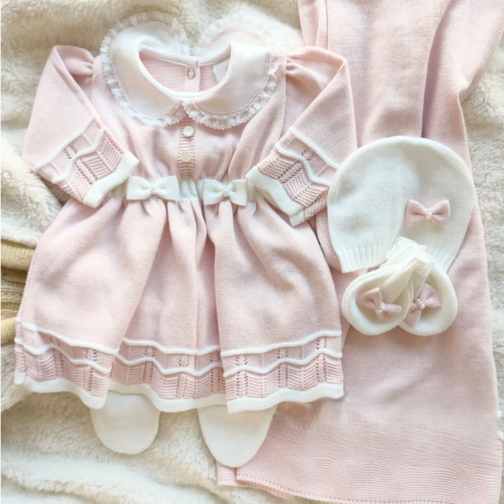 A baby clothing set laid flat on a soft cream blanket, featuring a pale pink long-sleeve dress with white lace trim and bow details, a matching pale pink bodysuit, a white bonnet, and white booties.
