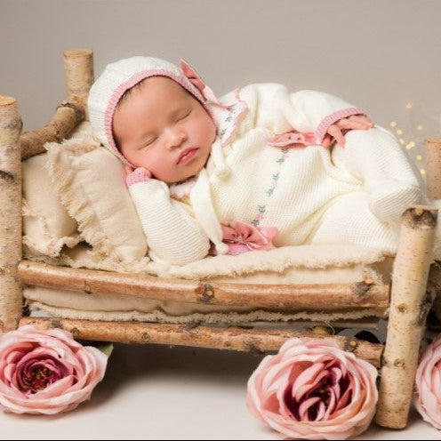 Newborn baby in a wooden crib with pink flowers on a neutral background Pink baby romper with floral details, newborn girl knit going home outfit with flowers embroidered details. the baby outfit is in pink. Baby girl knit coming home outfit pink and sweater in white, collar with handmade flowers