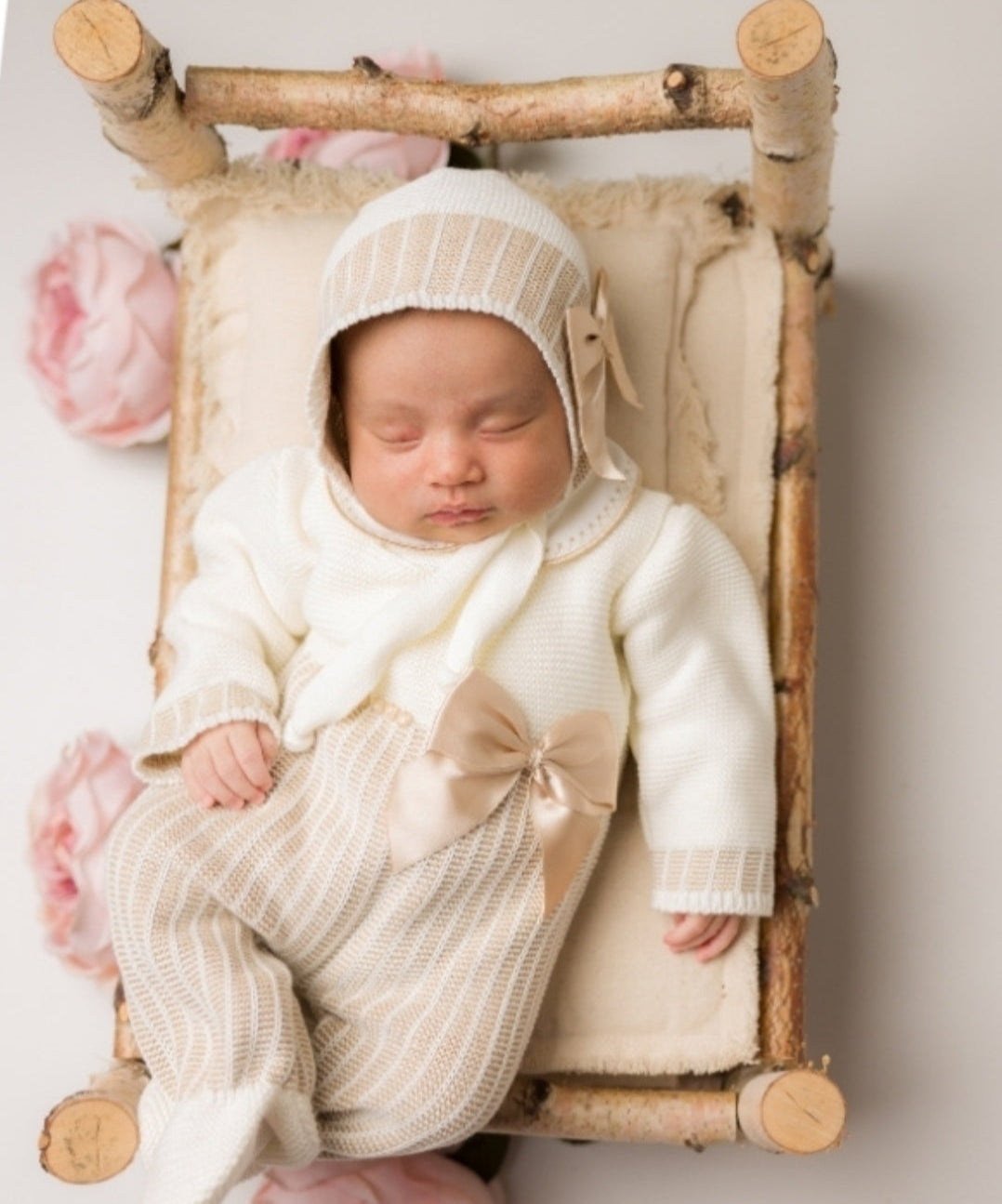 Newborn baby in a white outfit with a bow, lying on a small wooden bed with pink flowers.
