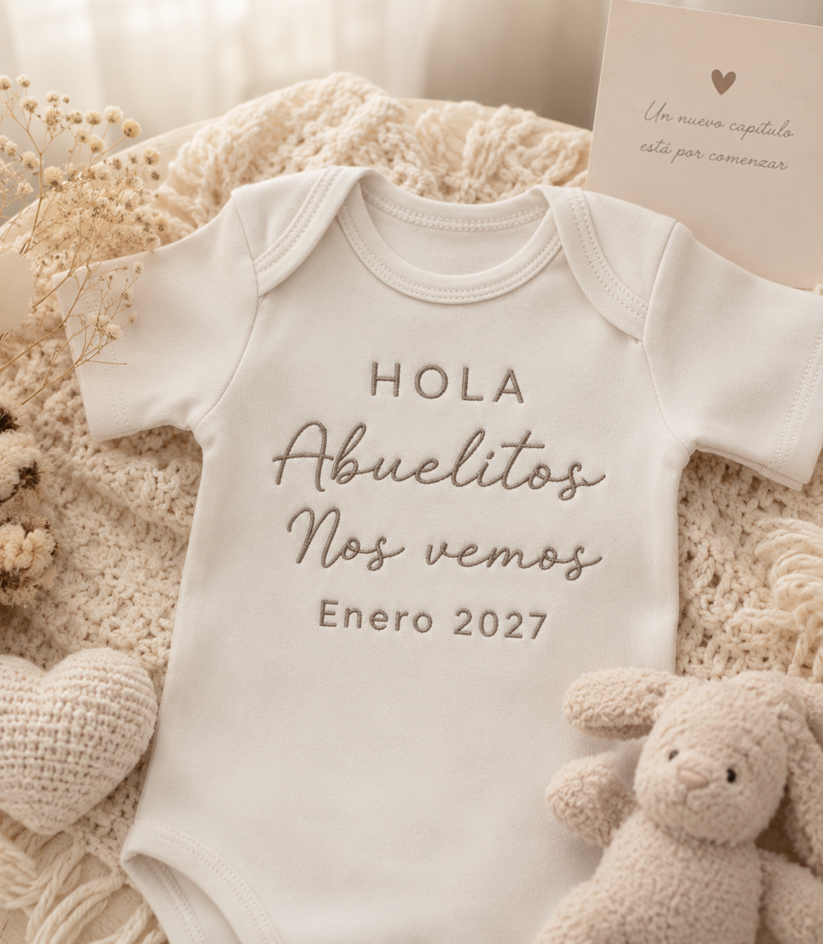 A white short-sleeve baby bodysuit laid flat on a cream knit blanket, printed with the Spanish text 'HOLA Abuelitos Nos vemos Enero 2027' in taupe lettering. A small card and plush toys are arranged around it as styled props.