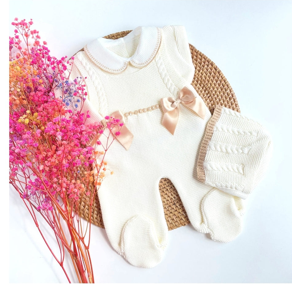 A knitted baby outfit in ivory and camel colors, consisting of a long-sleeve romper, a blanket, and socks, displayed alongside pink flowers.