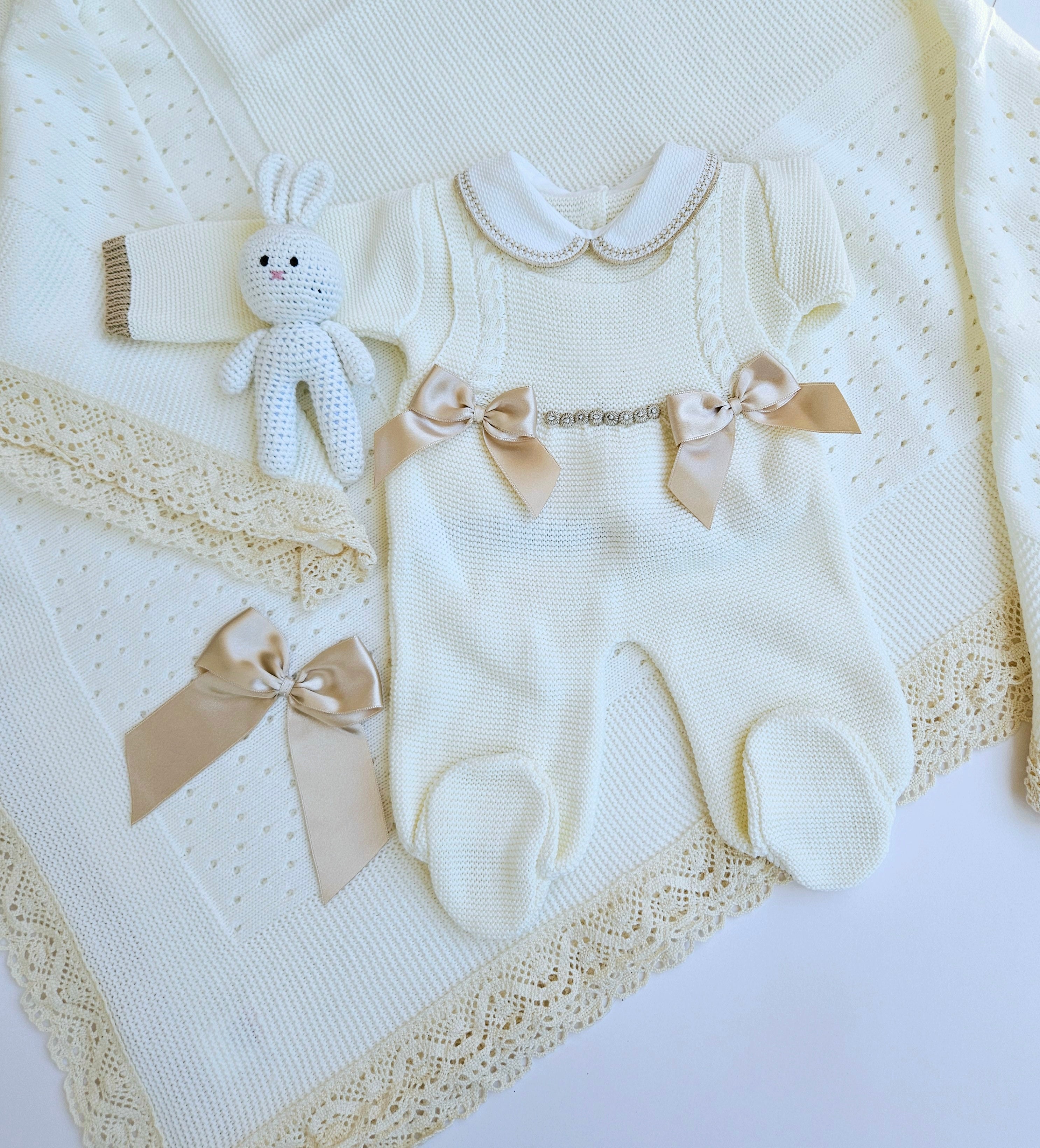 A knitted baby set in ivory and camel colors, including a romper, a blanket, and accessories, displayed on a white background.