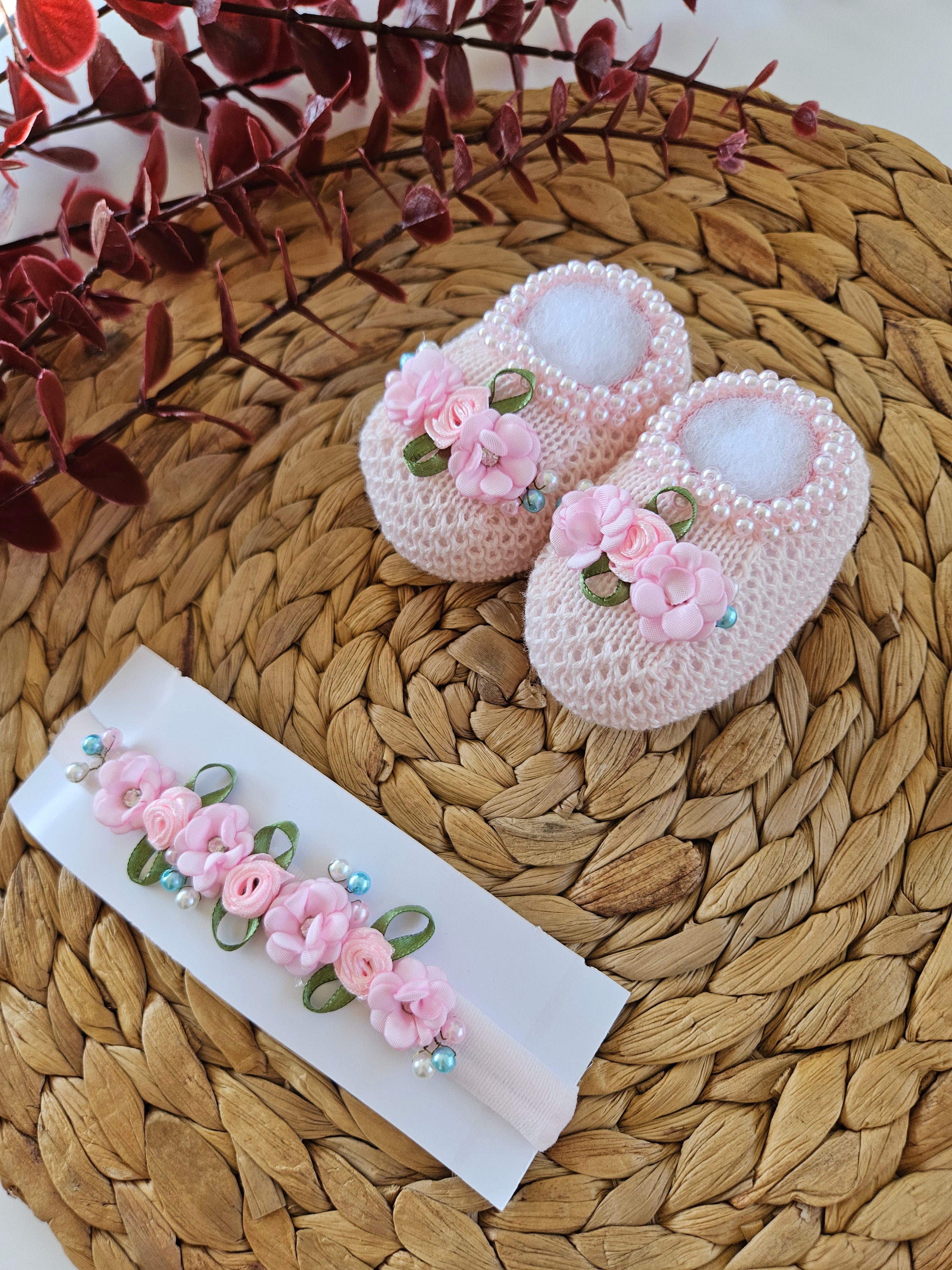 Handmade knitted pink baby bootie with floral pattern and a matching headband, displayed on a woven mat.