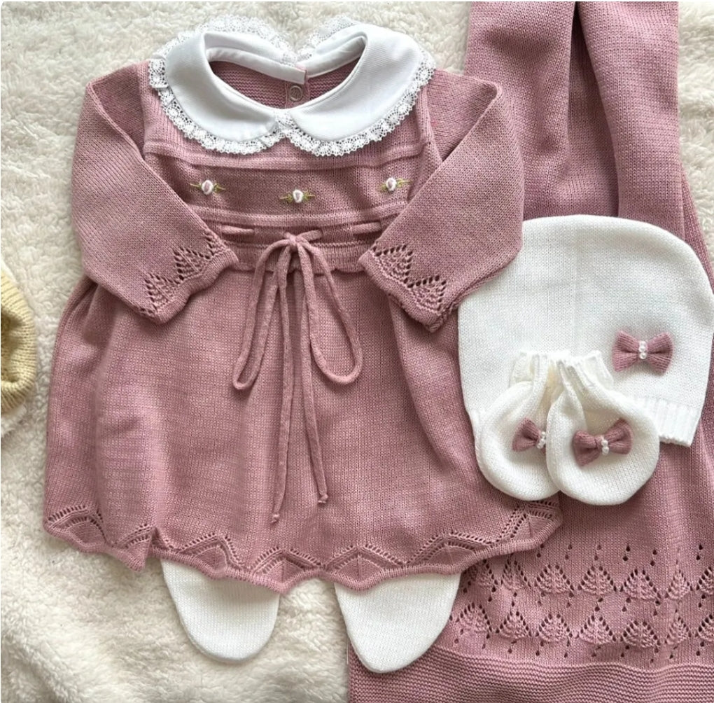 A pink knitted baby dress with embroidered flower details, accompanied by a matching hat and a pair of white baby mittens.