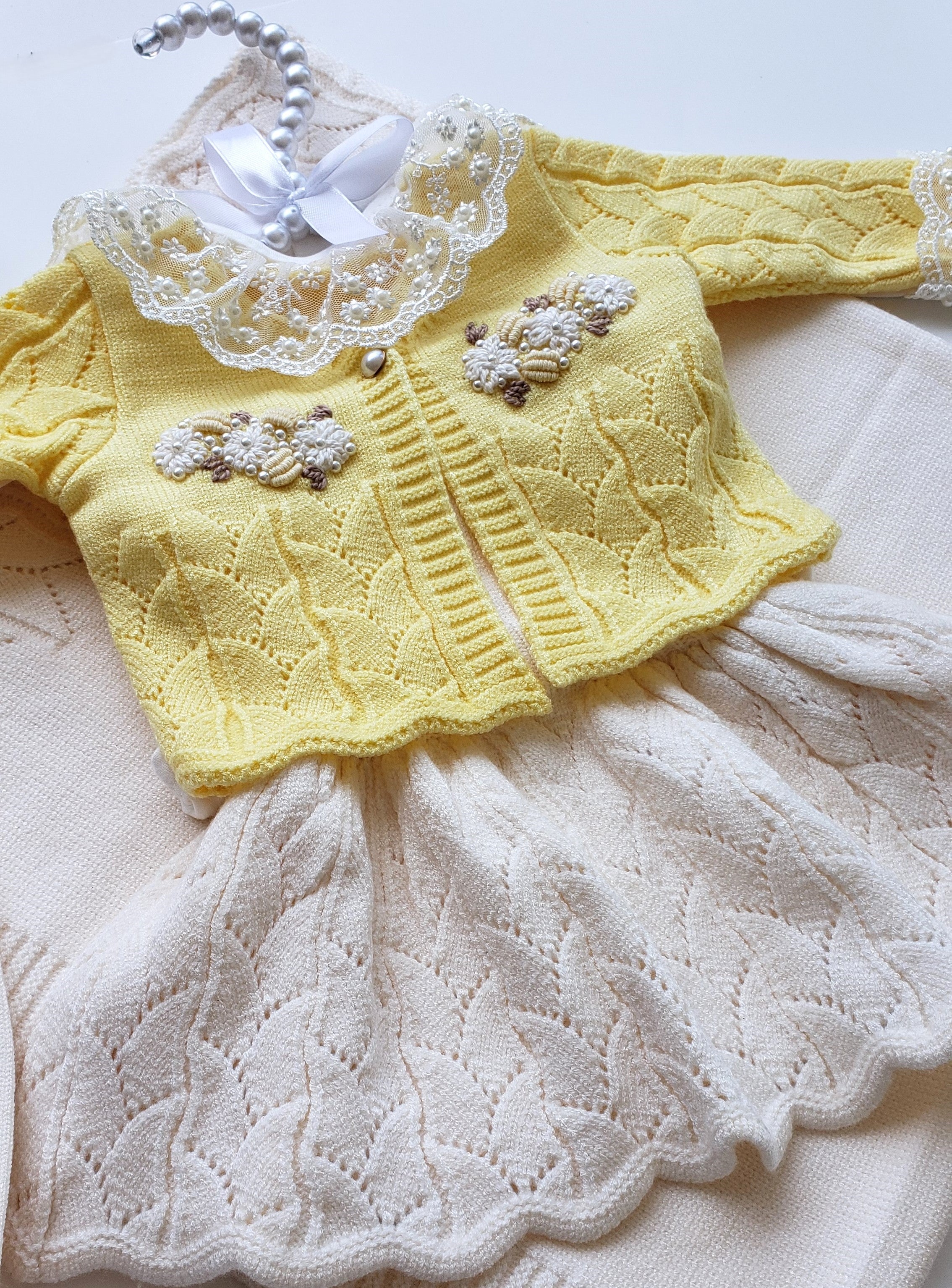 A baby knit outfit consisting of a pale yellow cardigan with a white lace collar and embroidered floral details, paired with an off-white knit dress or skirt, shown on a hanger against a light background.