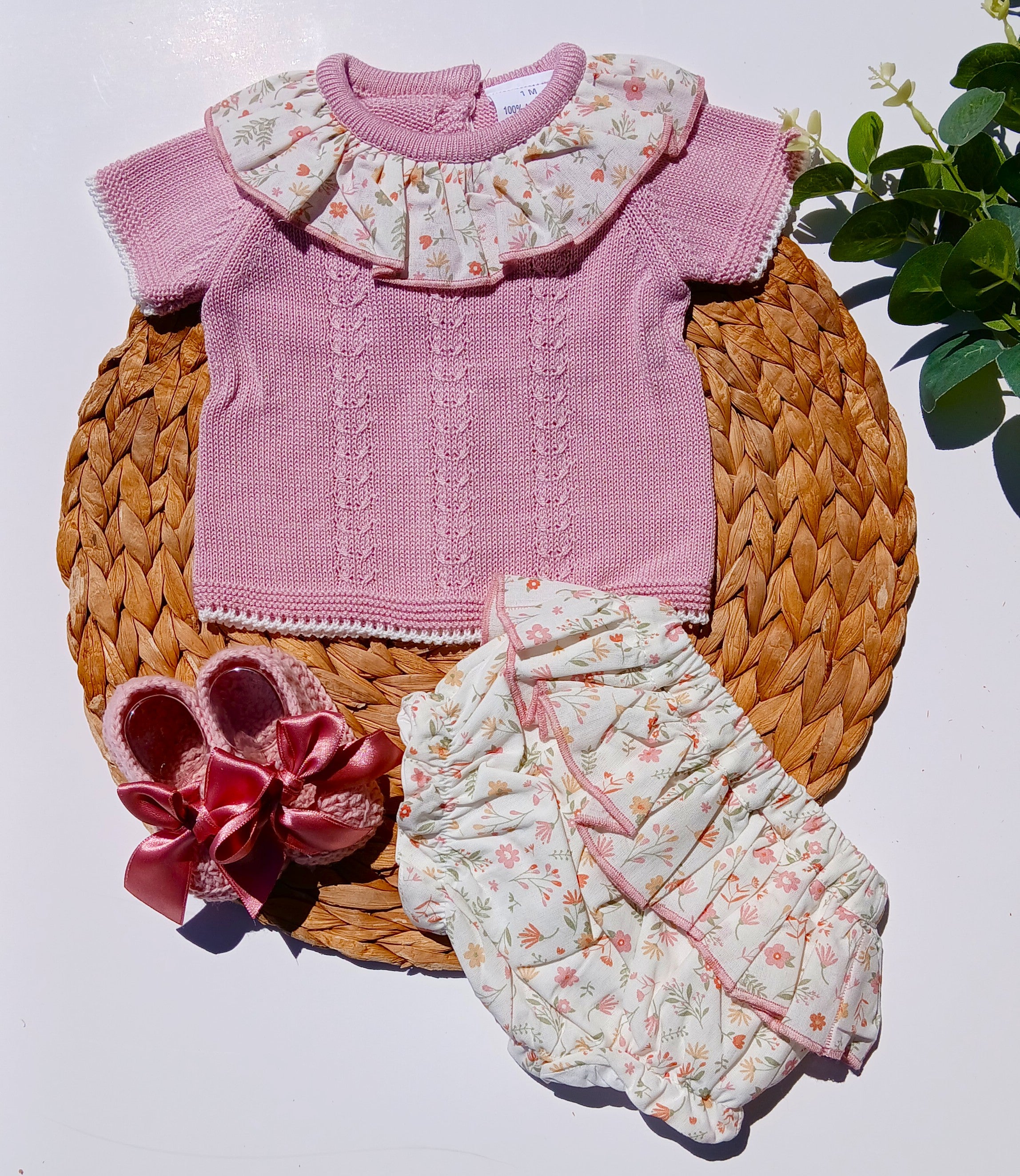 Baby outfit with pink knitted top, floral bloomers, and pink shoes on a woven mat.