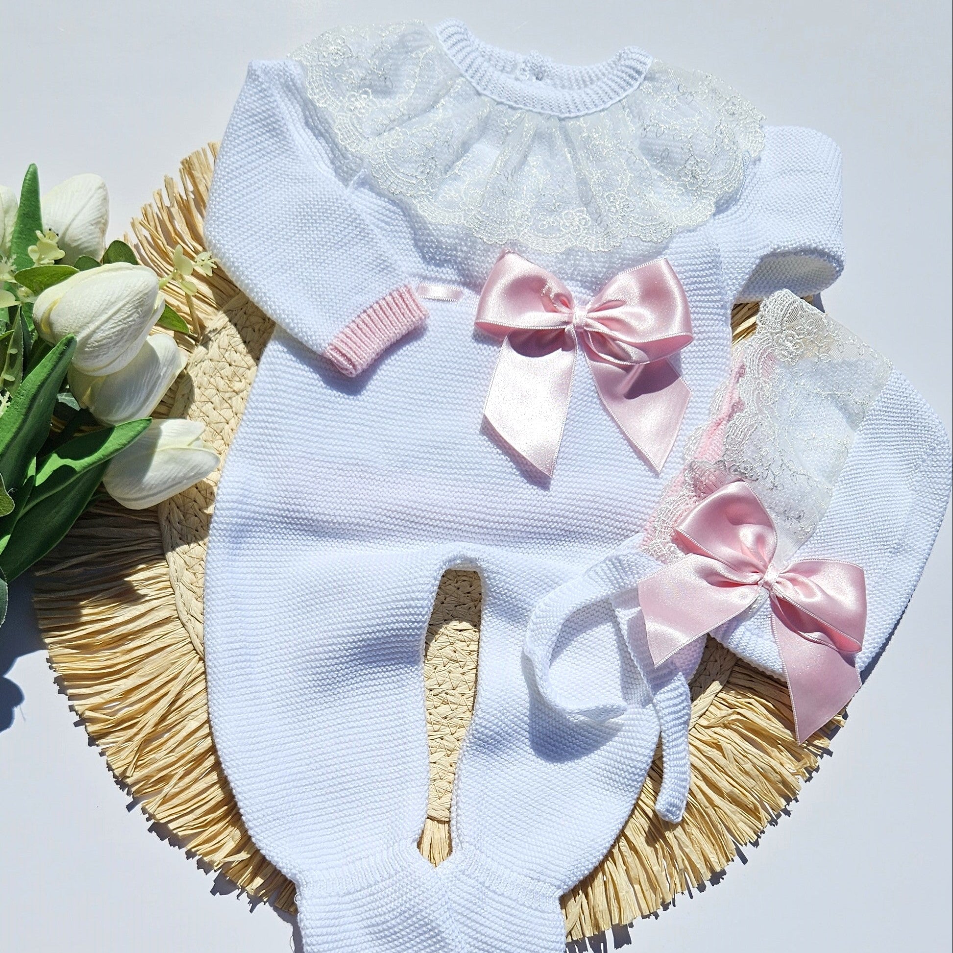 A baby knit romper in white with a lace ruffle collar, pink ribbed cuffs, and two large pink satin bows, displayed on a woven mat with white tulips.
