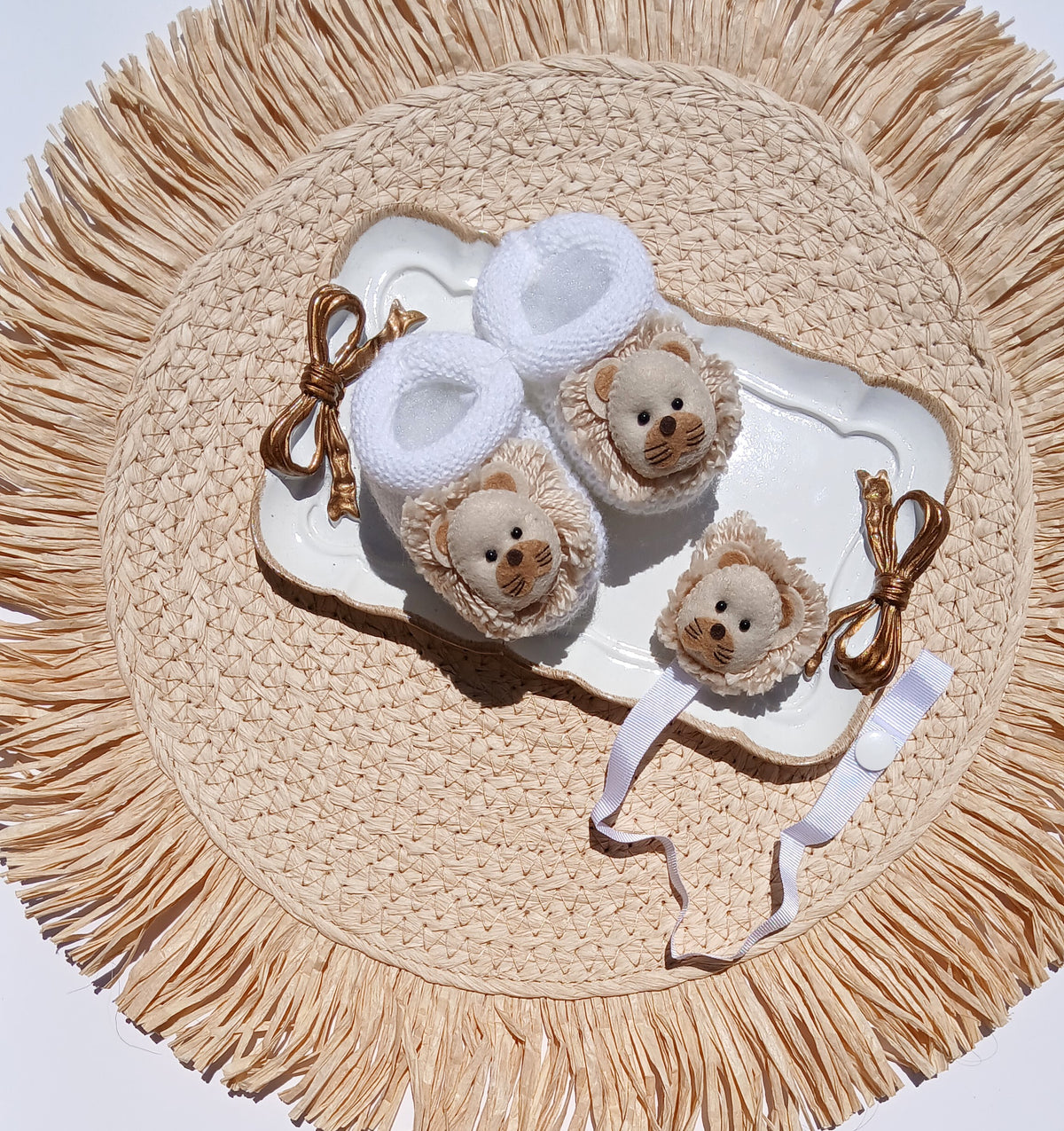 Three lion-face baby booties in white and beige knit with brown ribbon bows, arranged on a white tray over a round woven straw placemat.