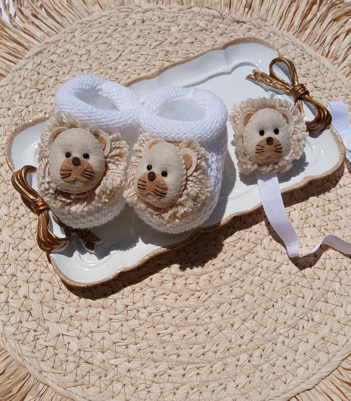 Three white and beige knitted lion-face baby booties arranged on a white scalloped tray, set on a woven straw mat. Lion pacifier chain 