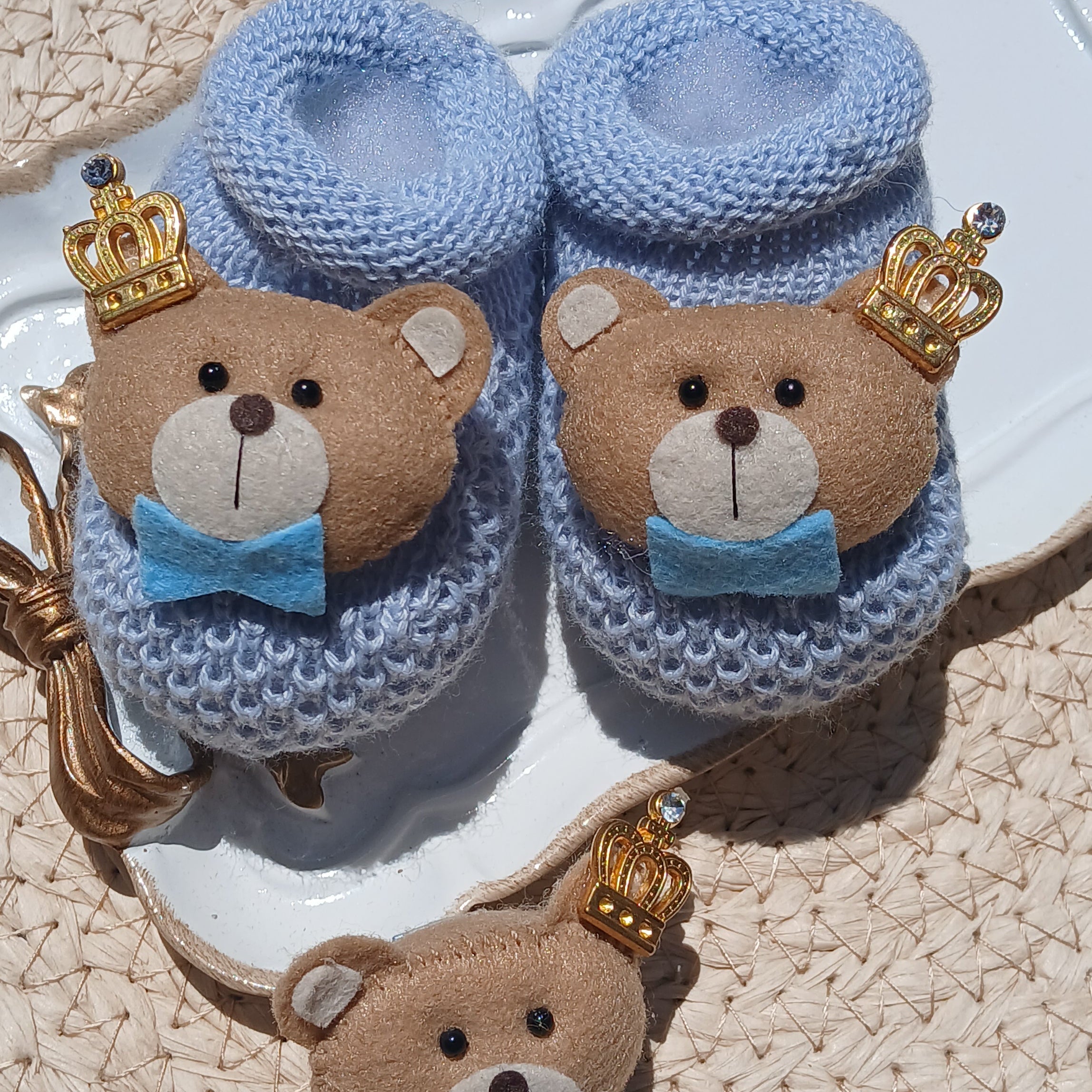 A pair of baby booties and a matching headband laid on a white tray. The booties are light blue knit with brown teddy bear faces, small gold crowns, and blue bows. Pacifier chain