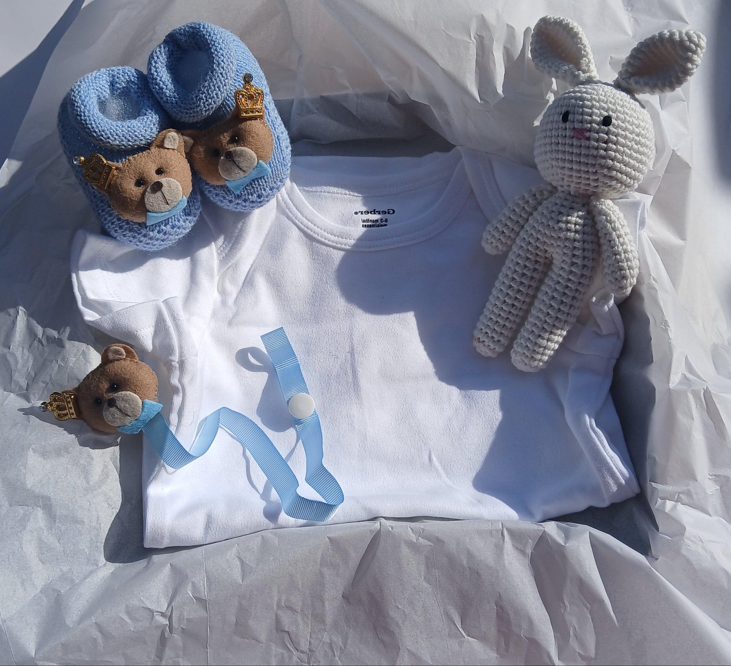 A baby gift set arranged on white tissue paper, featuring a white long-sleeve baby shirt, a small crocheted bunny toy, blue baby socks with teddy bear faces, and a matching blue ribbon.