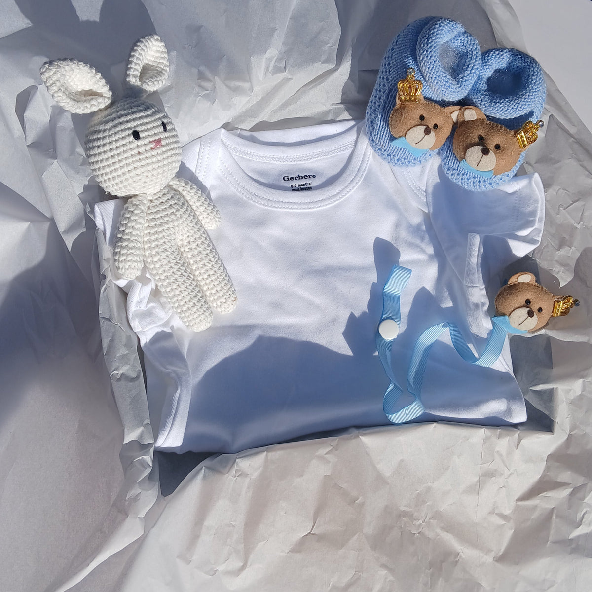 A folded white baby onesie displayed on crumpled white tissue paper, styled with a white crochet bunny toy, blue knit booties, and a blue pacifier clip.