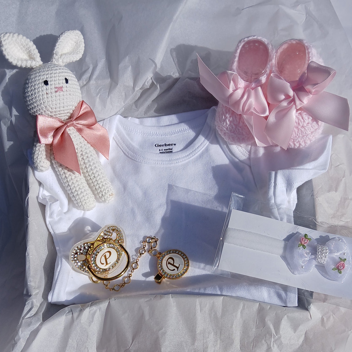 A baby gift set arranged on white tissue paper, including a white short-sleeve bodysuit, a pink knitted bunny headband with a bow, a white crocheted bunny toy with a pink bow, gold-tone jewelry, and a small packaged accessory.