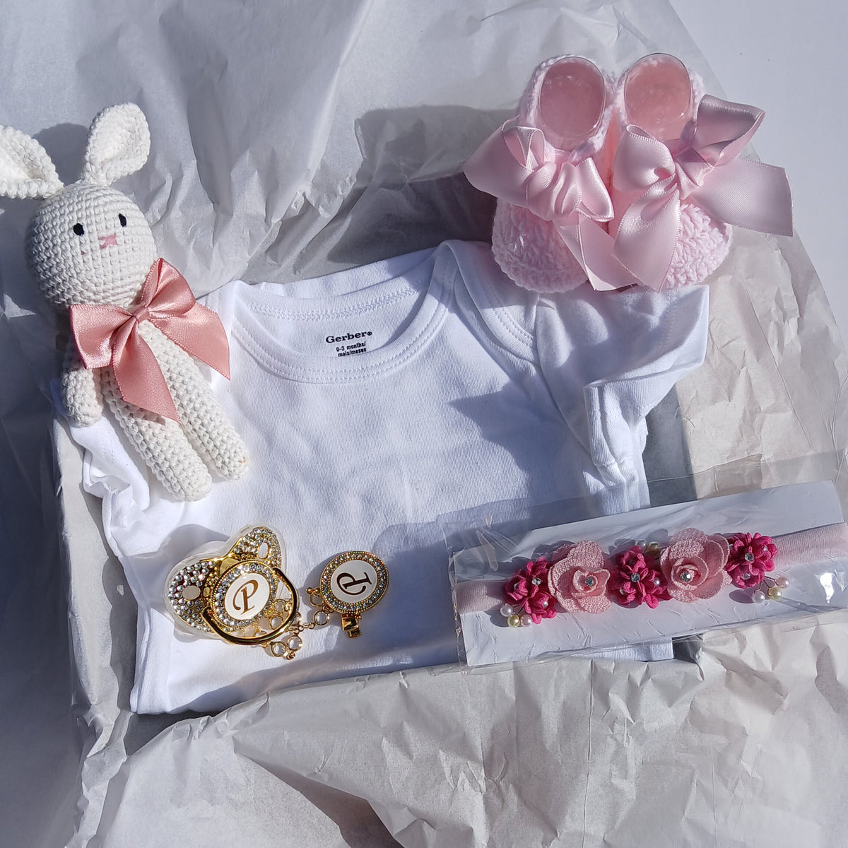 A baby gift set arranged on white tissue paper, including a white bodysuit, a pink bunny headband, a pink bow, a crocheted bunny toy with a pink bow, a gold heart-shaped brooch, and a pink floral hair clip.