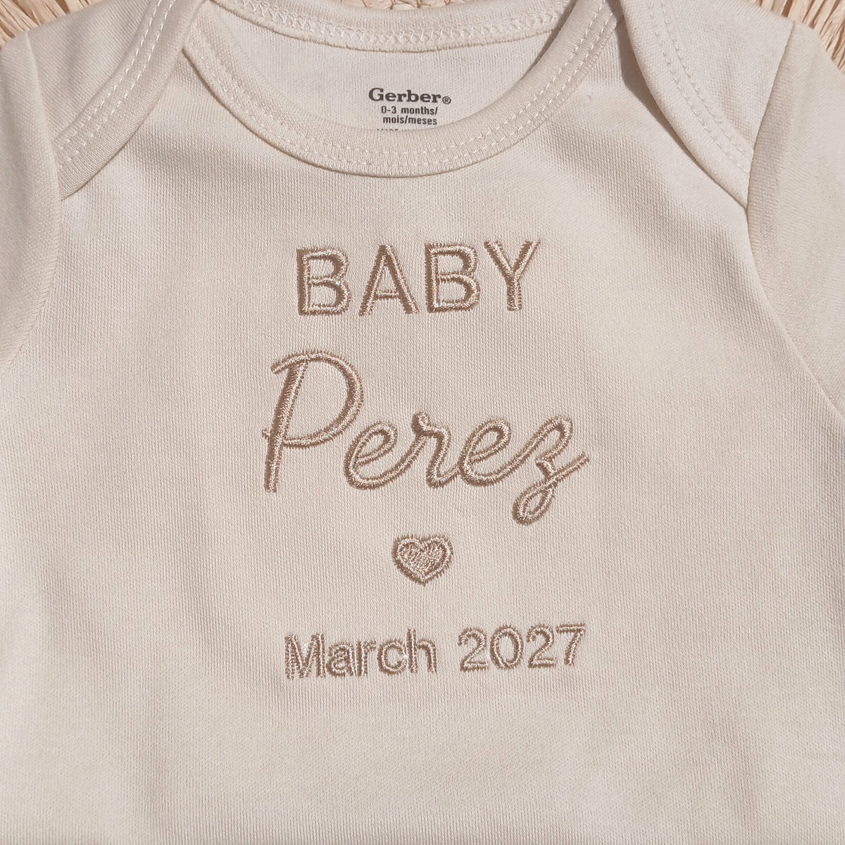 A close-up of a cream-colored baby bodysuit with 'BABY Perez March 2027' embroidered on the front, styled on a woven straw mat.