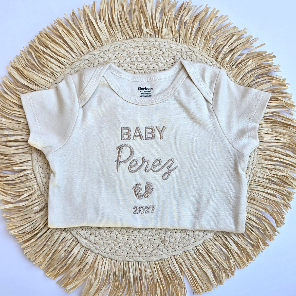 A short-sleeve baby bodysuit in cream/off-white with 'BABY Perez 2027' and footprints printed on the front, laid flat on a round woven straw mat against a light blue background.