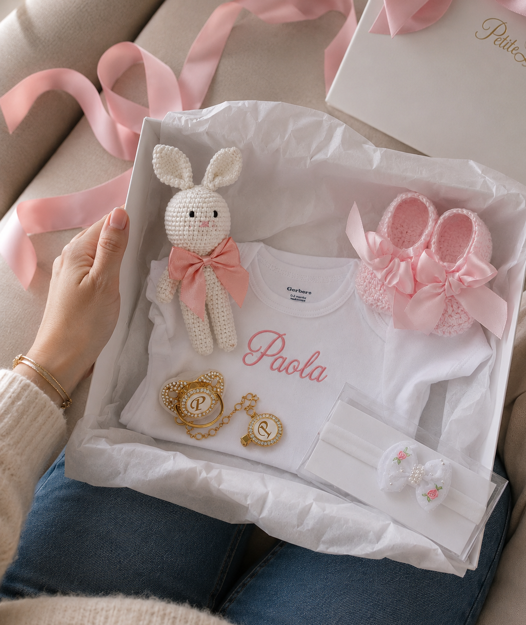 A baby gift set arranged in a white box on white tissue paper, held by a person. The set includes a white baby bodysuit with the name 'Paola' in pink script, a pink crocheted bunny toy with a pink bow, a pair of pink knit booties with bows, gold initial letter charms, and a white bow headband.